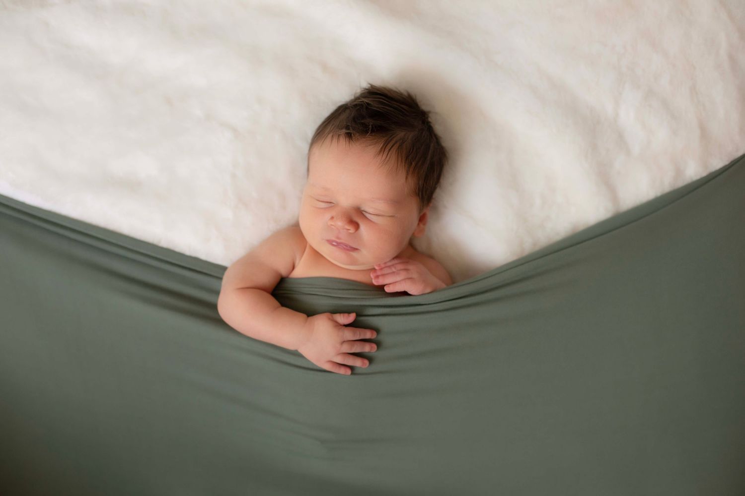Newborn baby sleeping peacefully wrapped in green fabric on white fluffy blanket in professional photo.