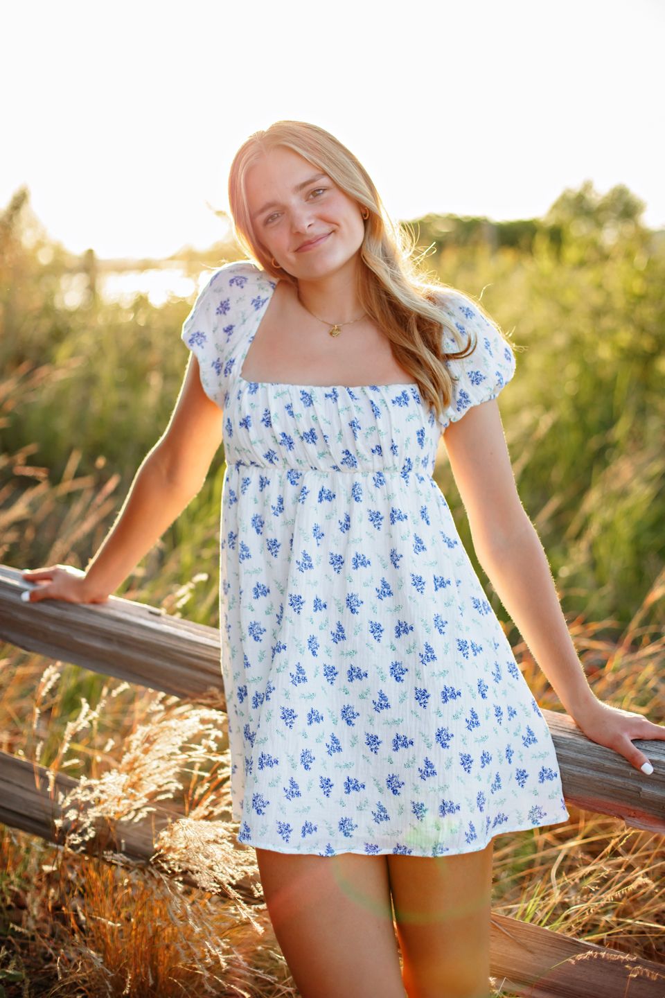 A summer dress with a delicate blue floral pattern glows warmly in golden sunset light as its wearer stands beside a railing.