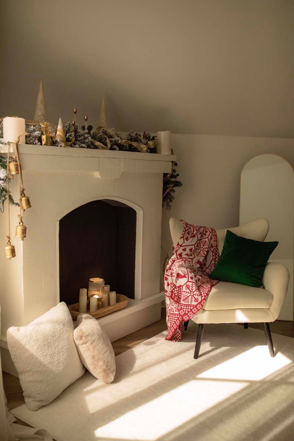 Cozy Christmas living room with white fireplace, candles, festive decor, cream armchair with red blanket and green pillow.