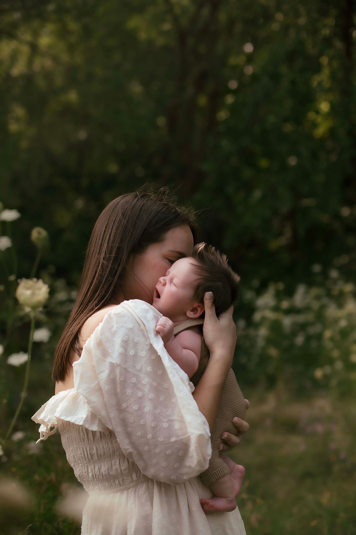 A mother in a white dress tenderly kisses her sleeping newborn baby outdoors among wildflowers.