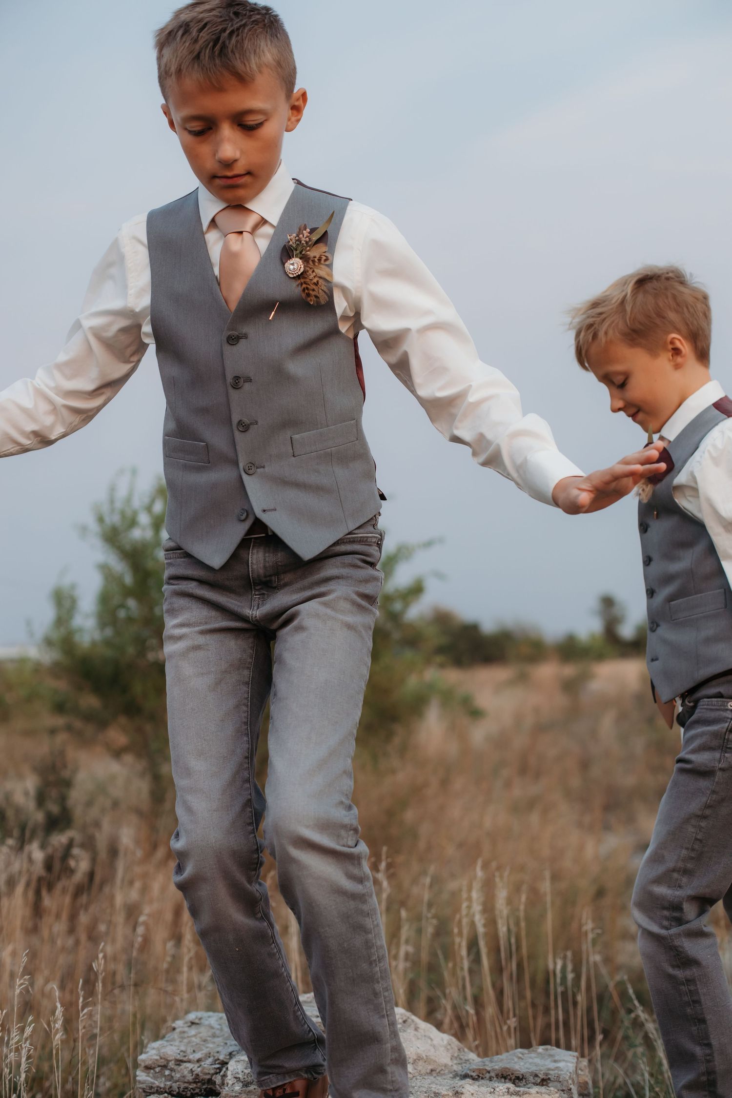 Two boys in matching grey vests and formal wear playing on rocks in field.