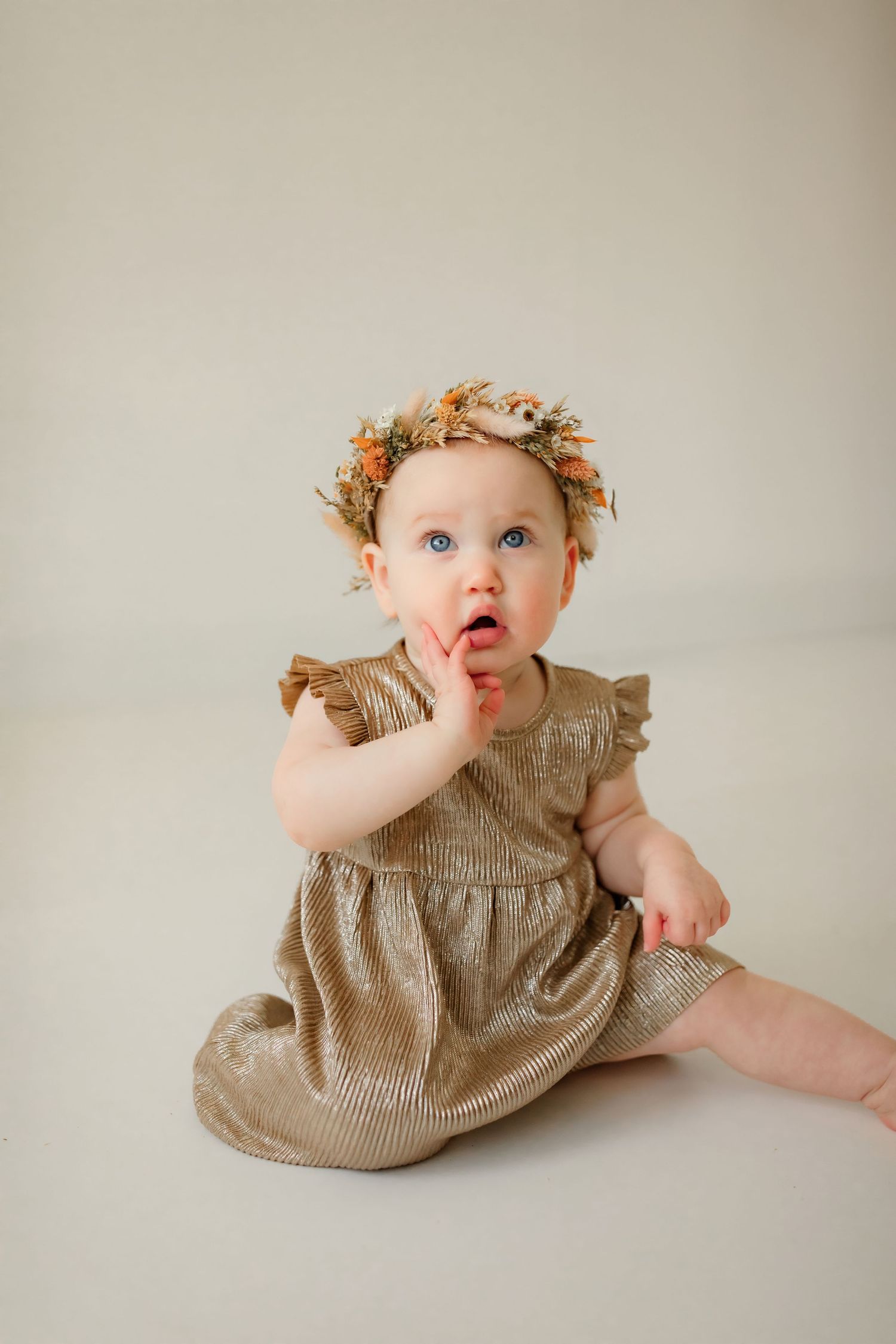Baby in beige dress with curly hair sits on white background.