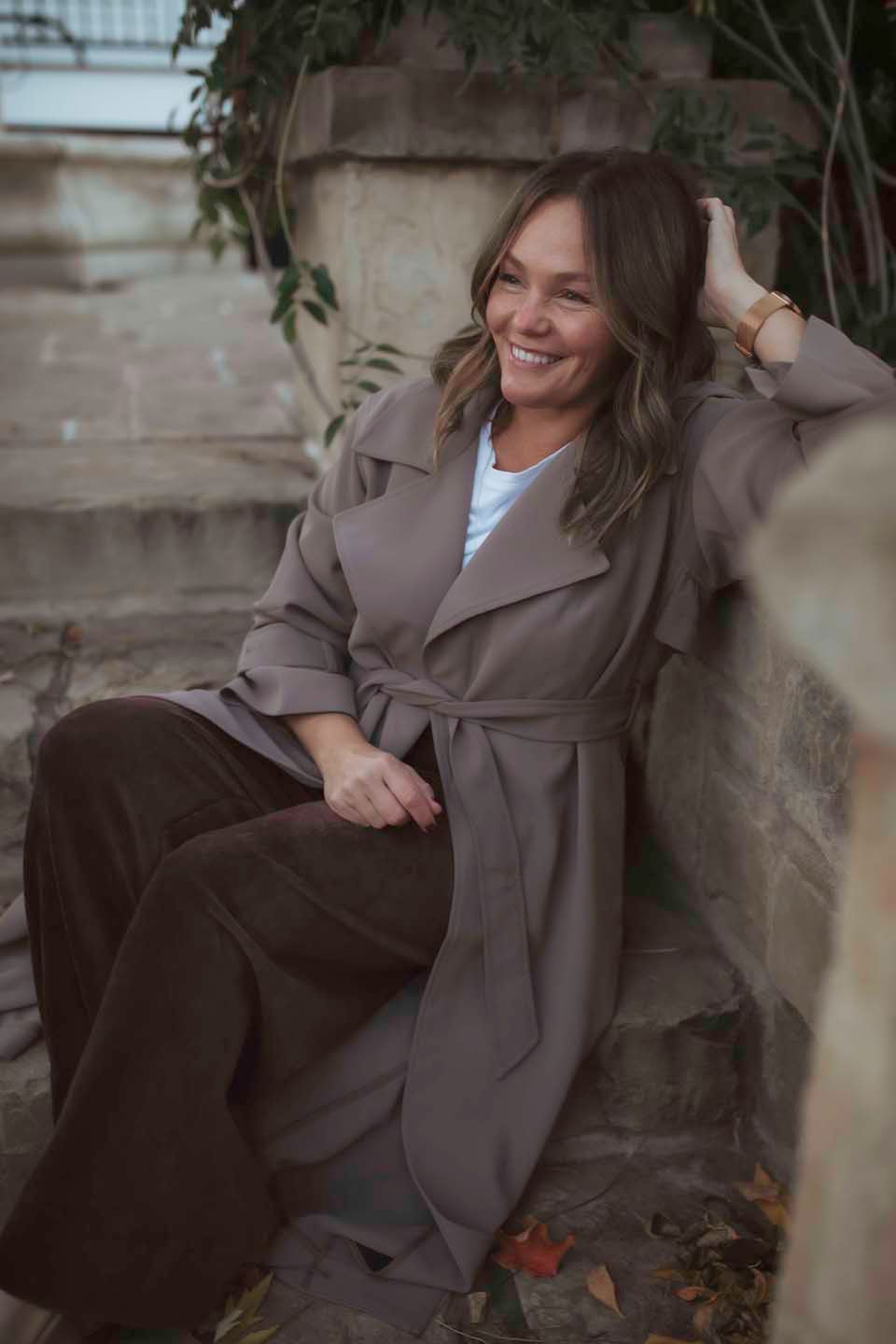 Person in brown trench coat laughing while sitting outdoors.