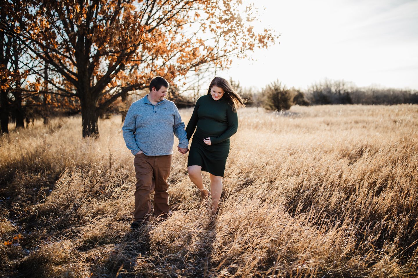 A couple walks together through a golden autumn field with dried grass and fall foliage in the background.