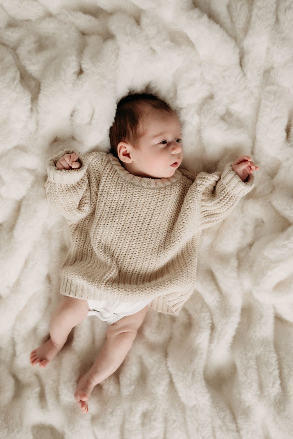 Baby in oversized knit sweater stretches out on fluffy white bedding.