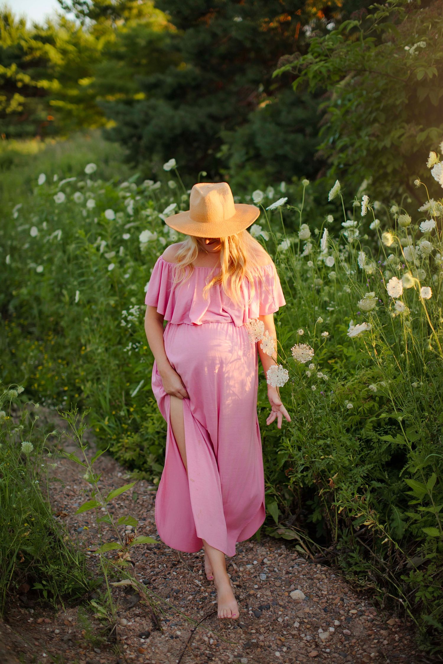 Pregnant woman in pink dress and yellow hat walks barefoot on garden path surrounded by wildflowers in golden sunlight.