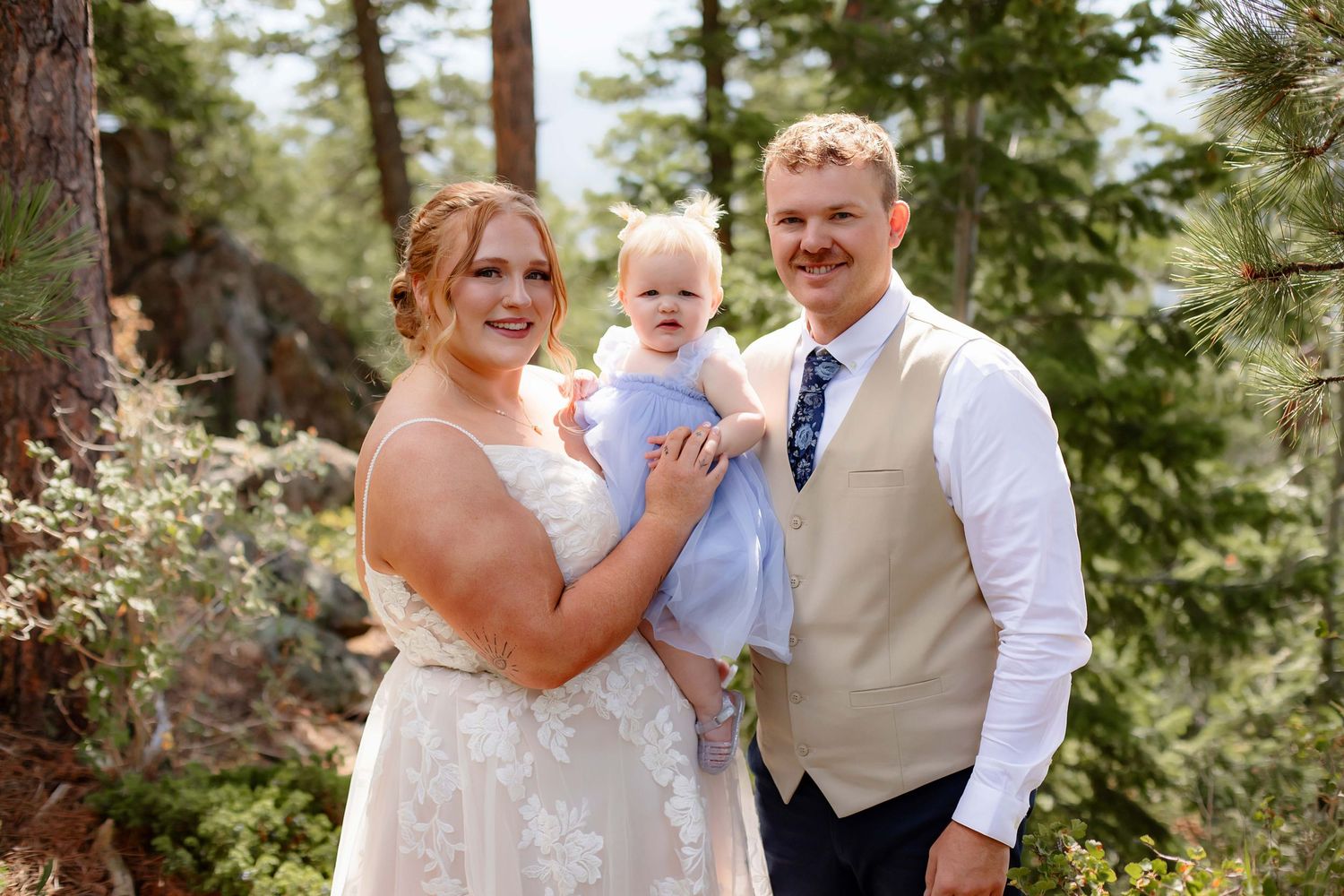 A wedding couple and baby pose together outdoors in a scenic forest setting with pine trees in the background.