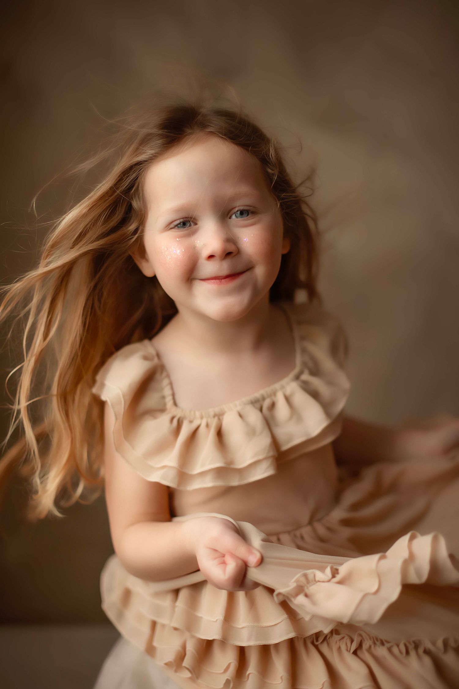 A series of joyful portraits showing a young child in a beige ruffled dress twirling and playing against a brown backdrop.