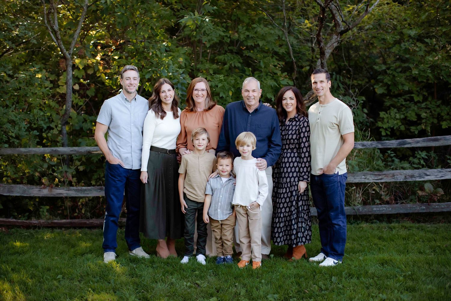 Extended family gathering poses for outdoor portrait in front of rustic wooden fence.