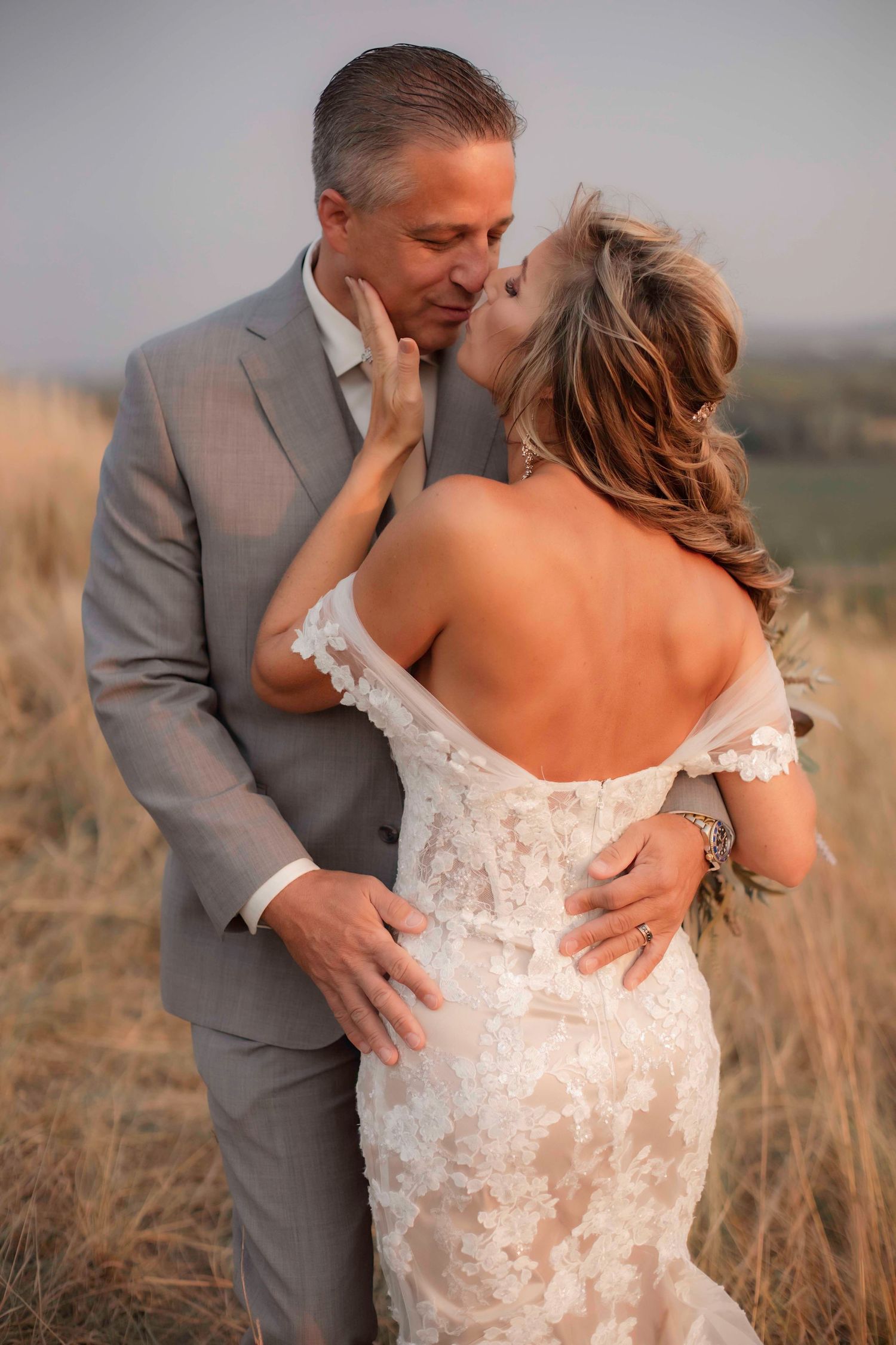 Tender moment between newlyweds in golden field with bride in lace dress and groom in grey suit.