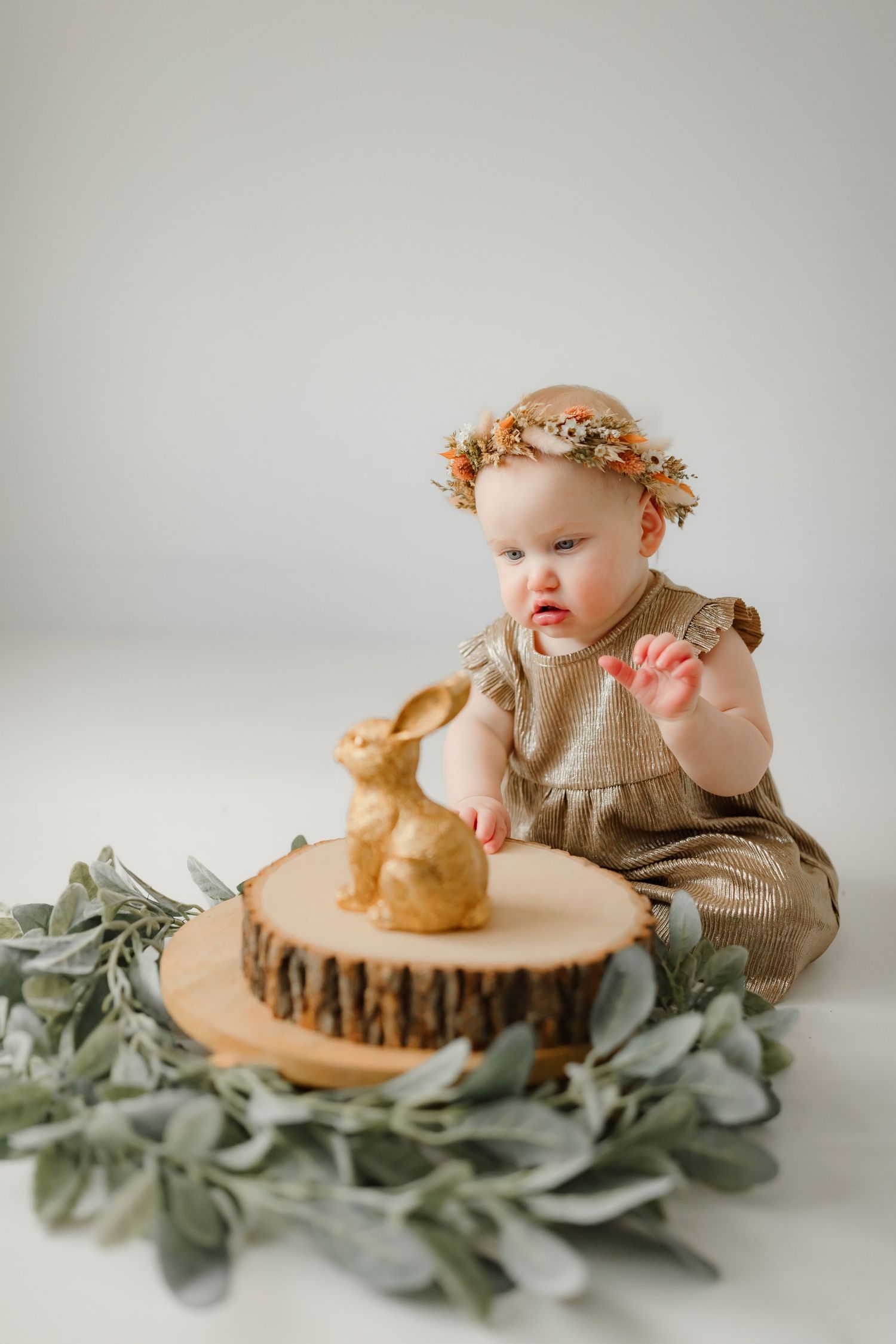 Baby in knit outfit with flower crown looking at bunny decoration on wooden slice.