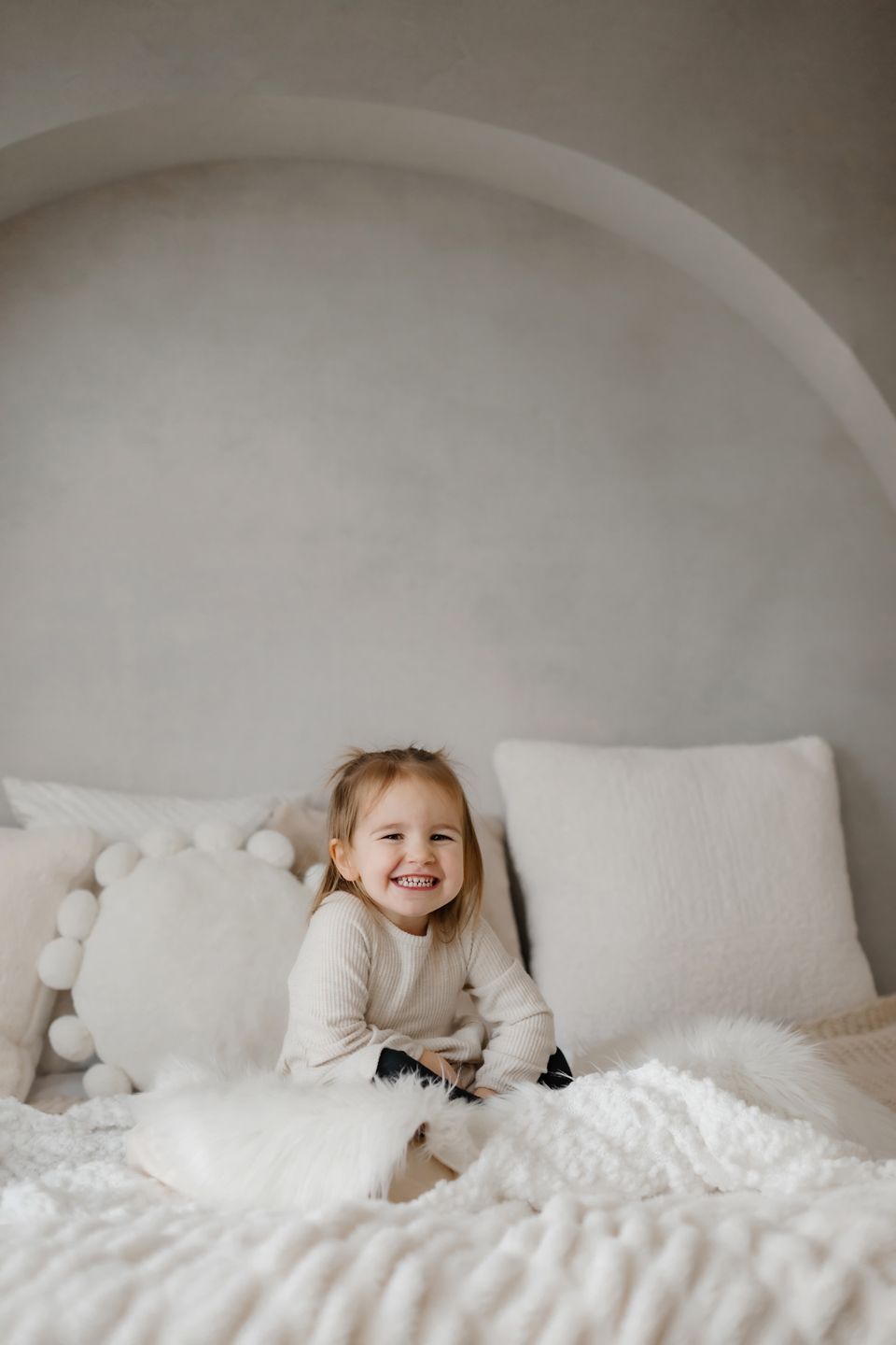 A child in a cream sweater sits happily among white pillows in an arched bedroom nook.