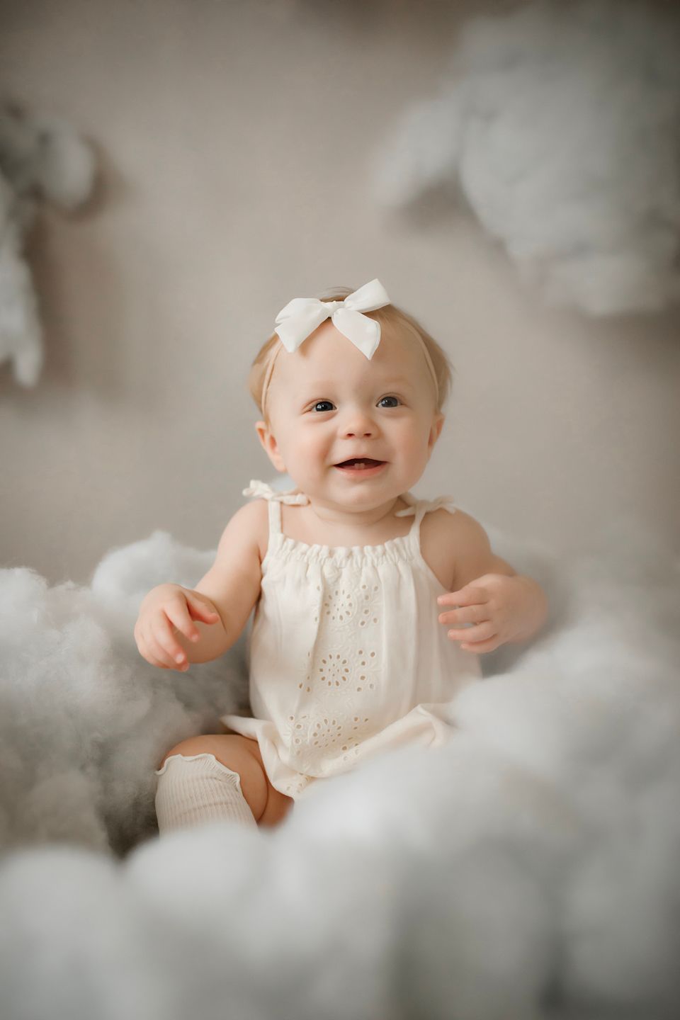 Baby in white dress sitting among fluffy cloud decorations with joyful expression.