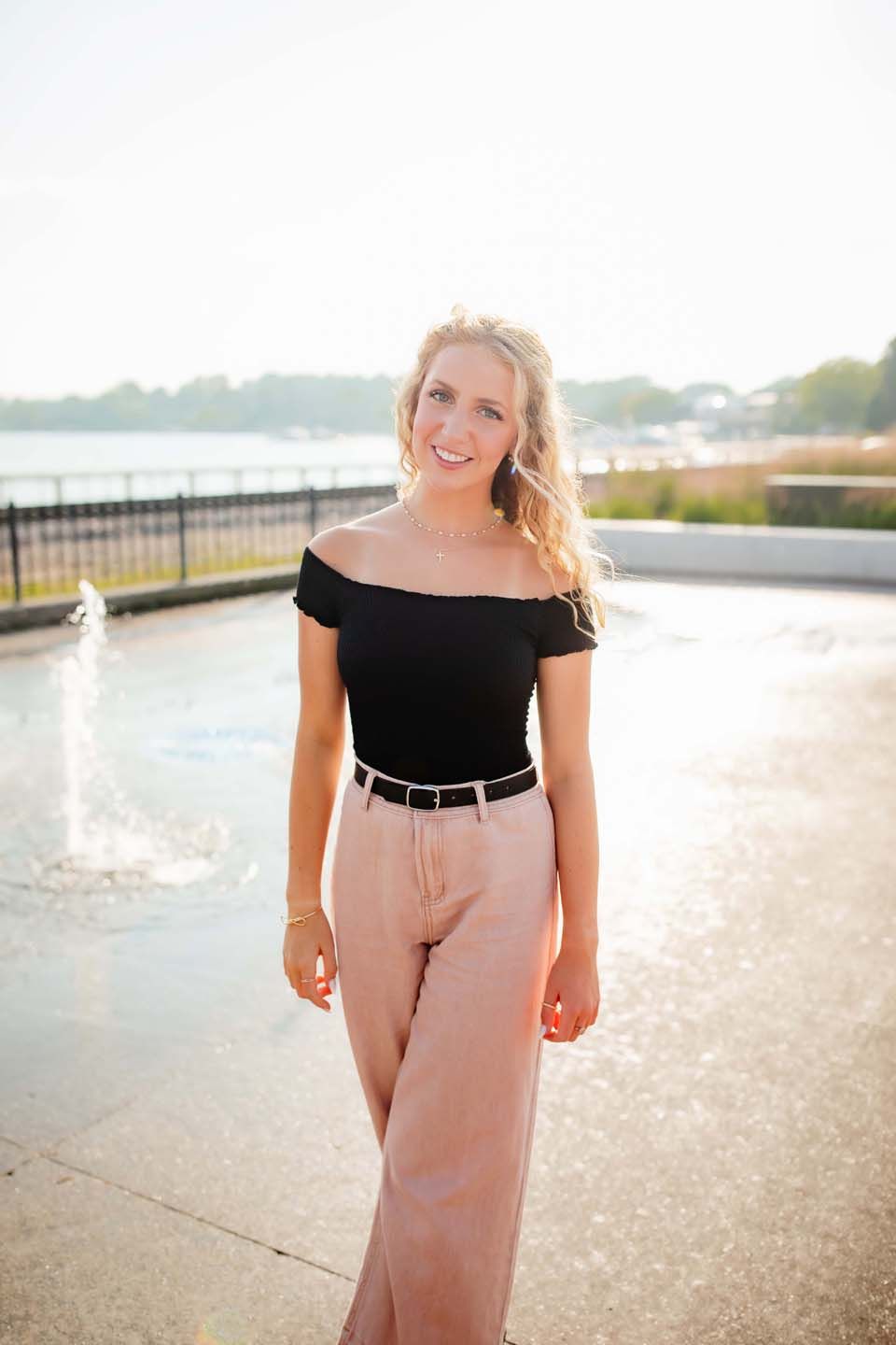 Standing by a fountain wearing an off-shoulder black top and blush wide-leg pants.