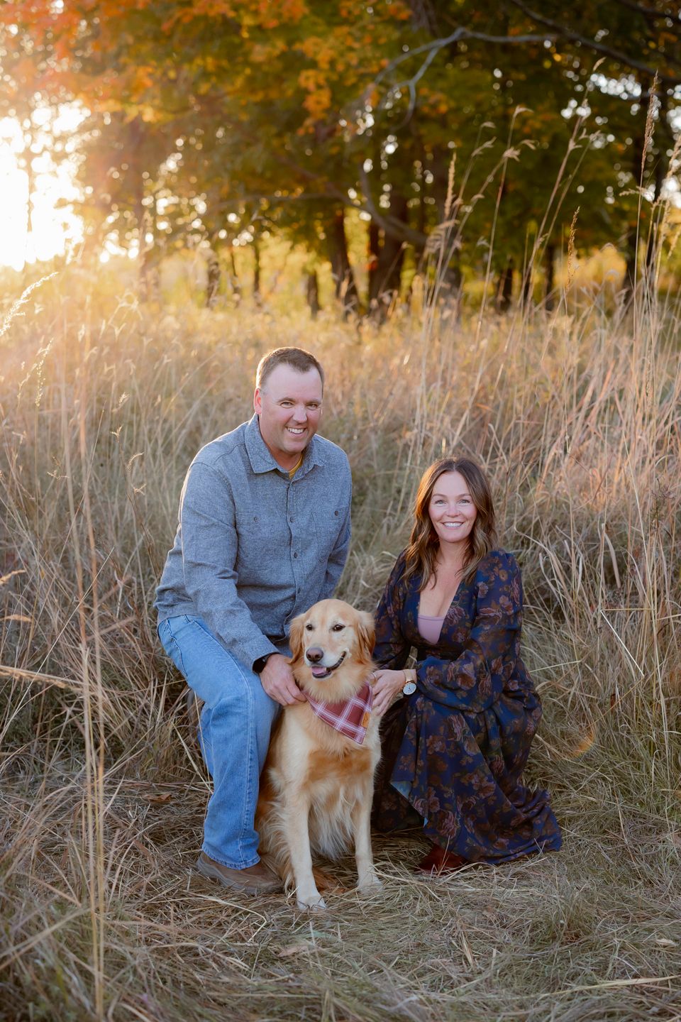 Golden retriever with two people posing in tall grass during sunset.