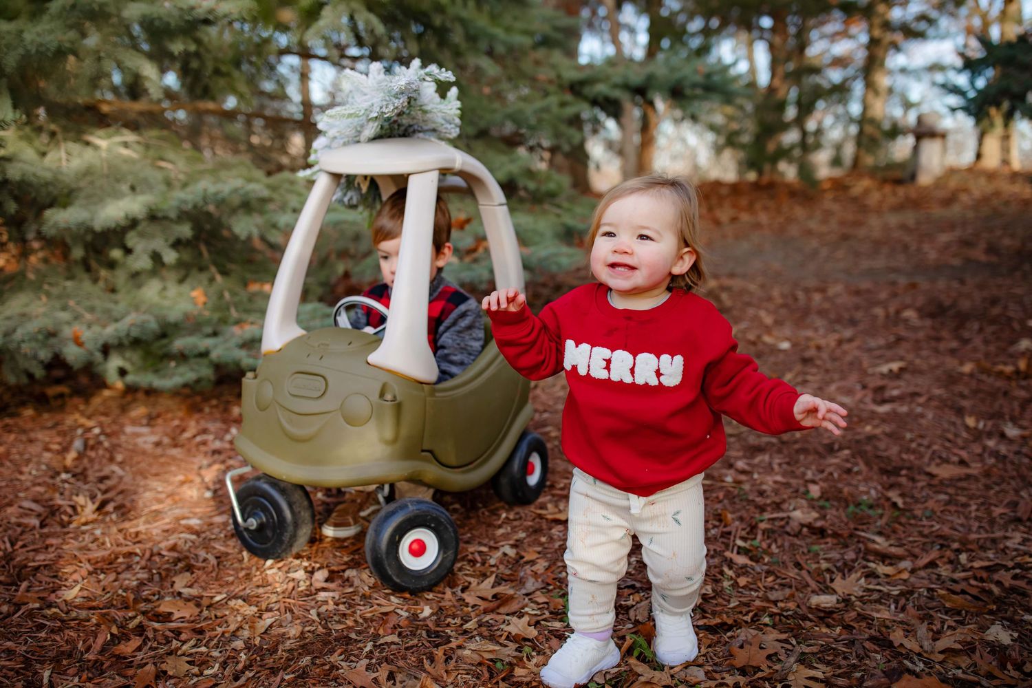 Young child in red sweater stands next to decorated toy cozy coupe car on forest path.