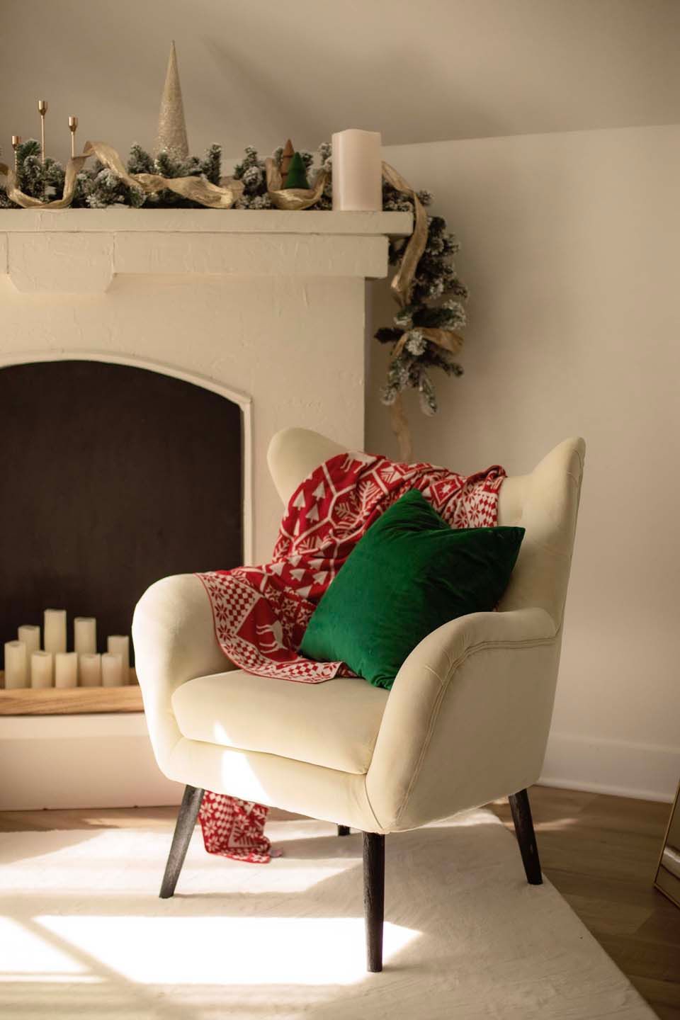 Cozy cream armchair with green velvet pillow and red blanket beside a Christmas-decorated fireplace with candles.