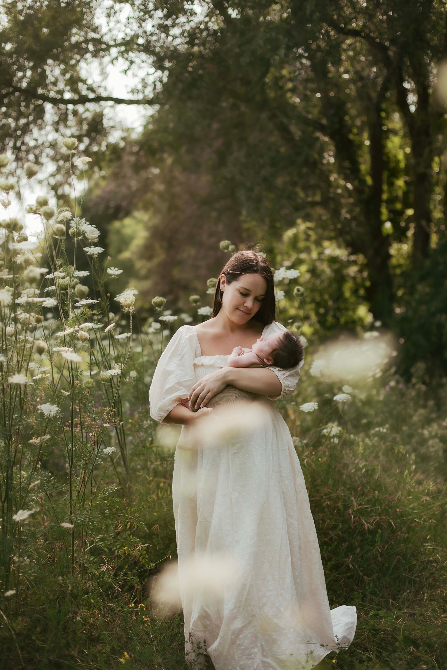 Woman in elegant white gown holding sleeping newborn in a field of white wildflowers.
