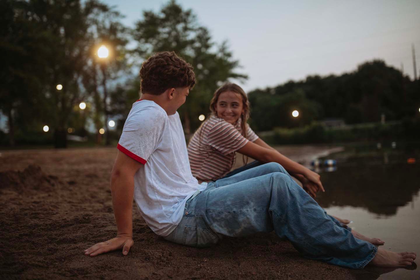 Two young people sit on a sandy riverbank at dusk, sharing a conversation in a park setting.
