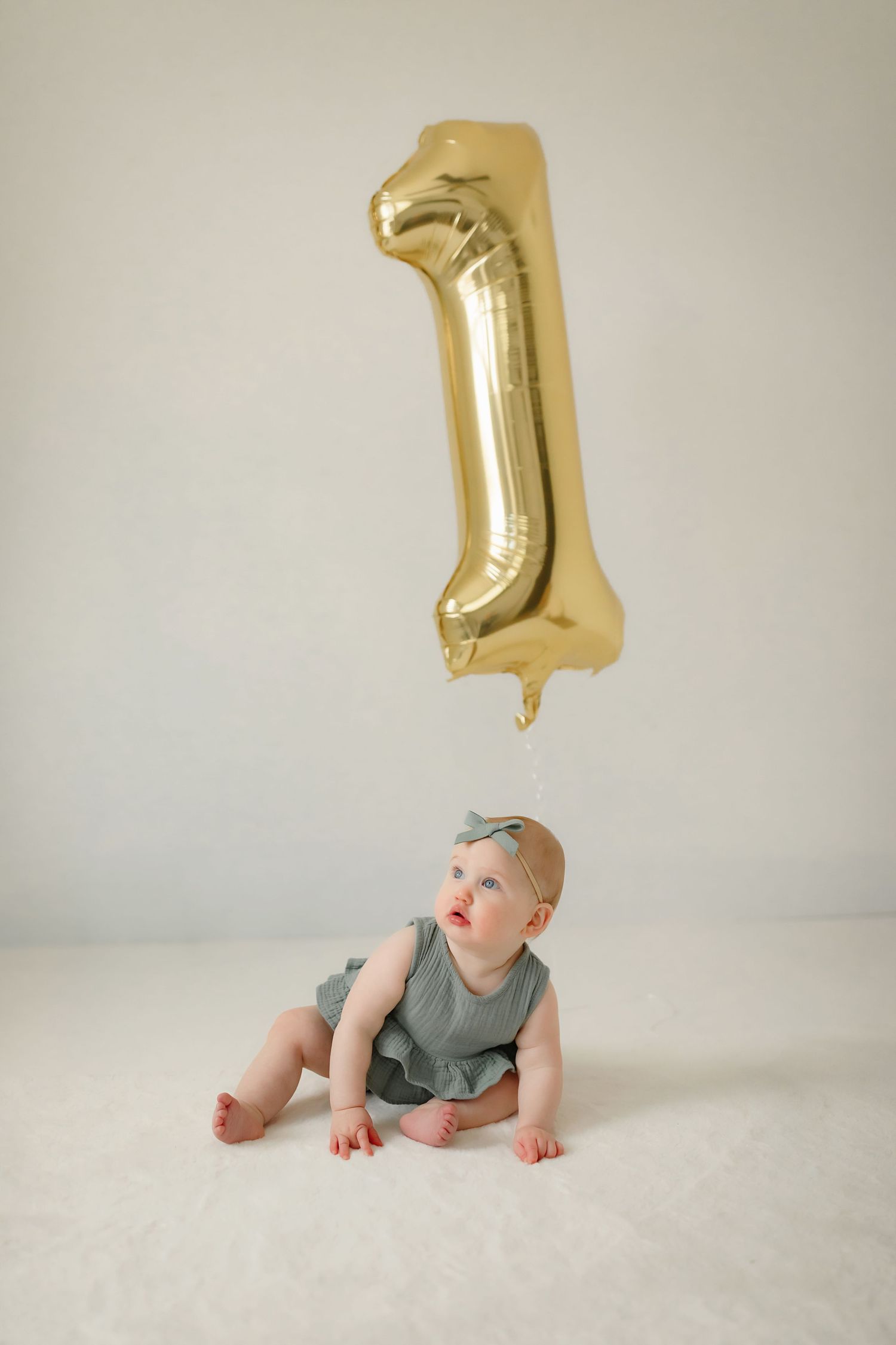Baby in grey romper sitting under gold number one balloon against white background.
