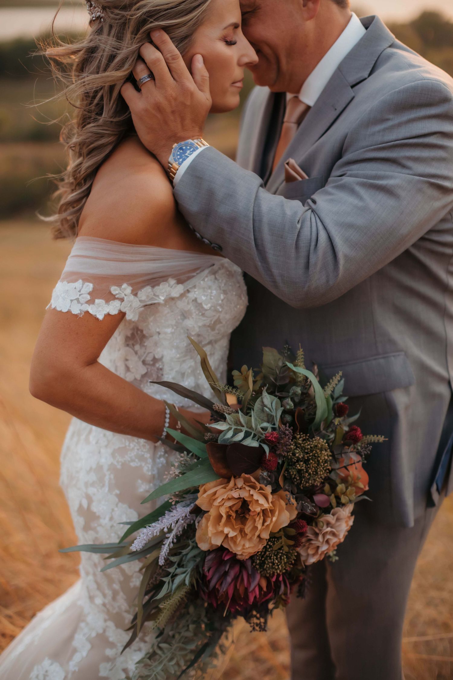 Romantic wedding portrait with bride holding autumn bouquet while groom in grey suit embraces her.