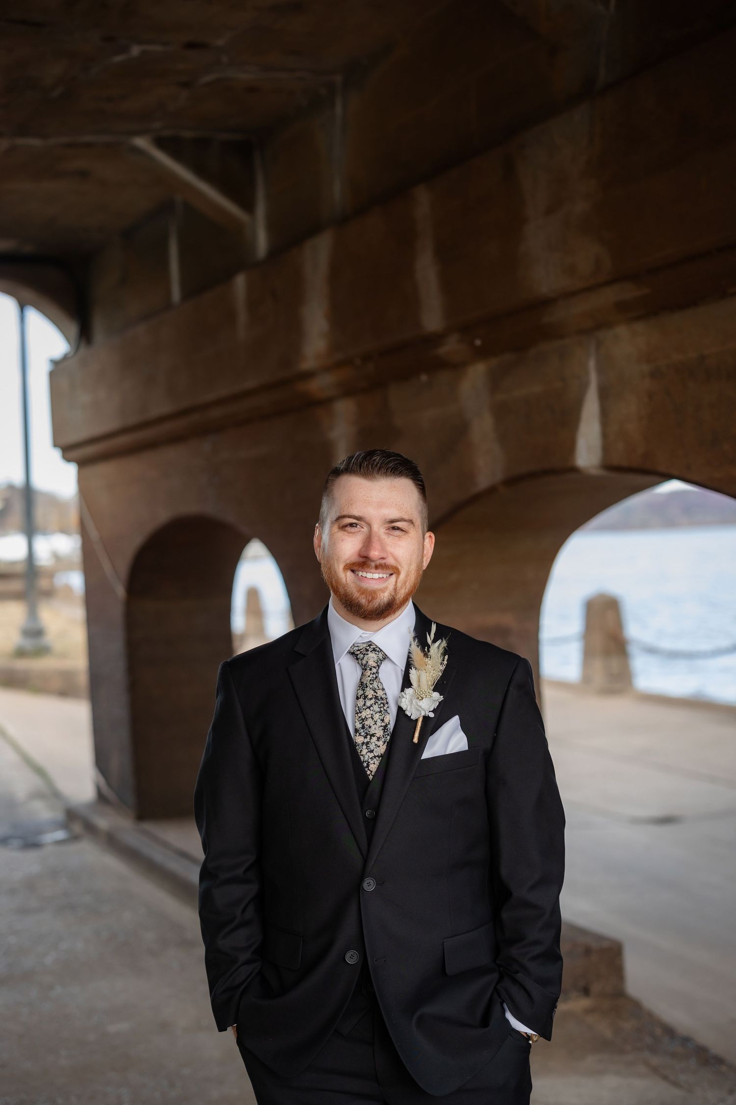 A groom in a black suit with floral tie and boutonniere poses under an archway.