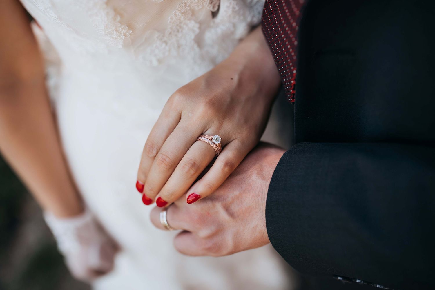 Close-up of wedding rings and clasped hands showing red nail polish and formal attire.