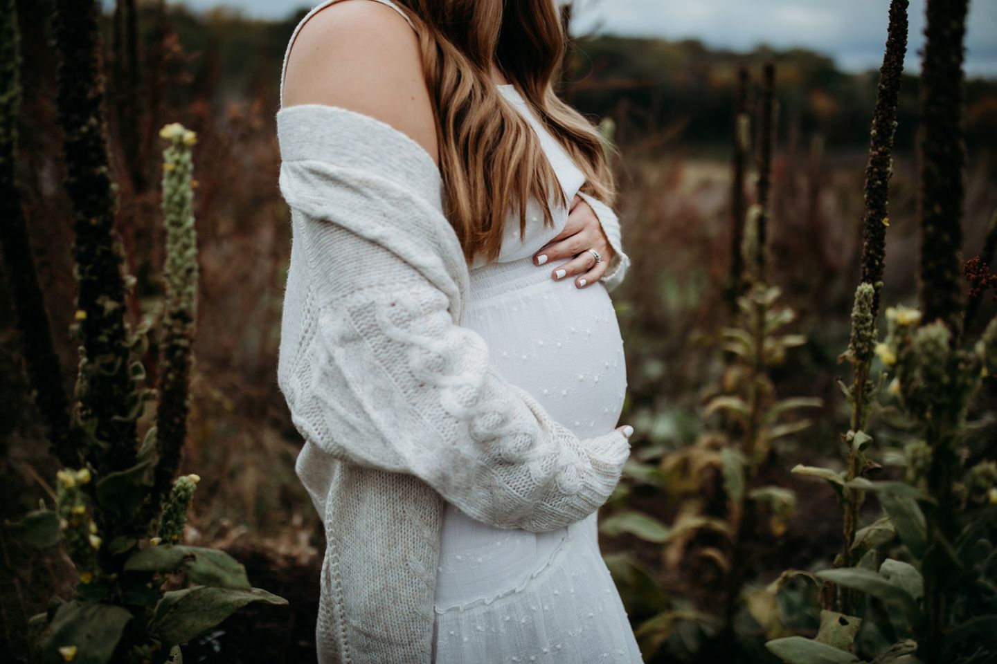 Expectant mother in a white dress cradles her baby bump in a garden setting.