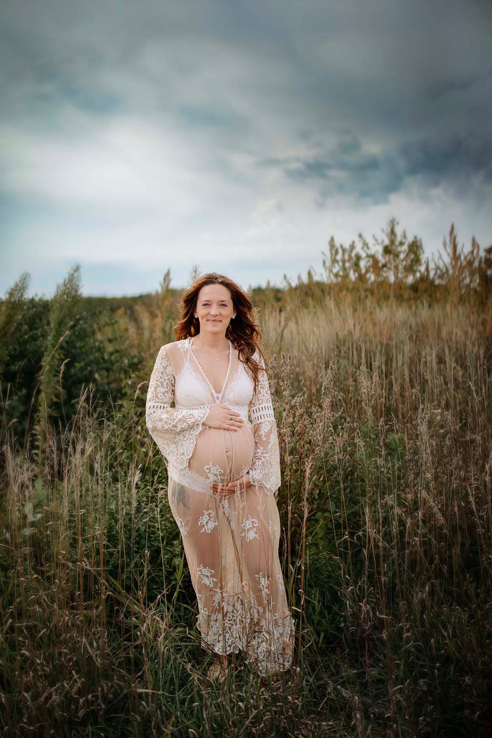 A maternity photo taken in a field of tall grass under cloudy skies features an elegant lace dress.