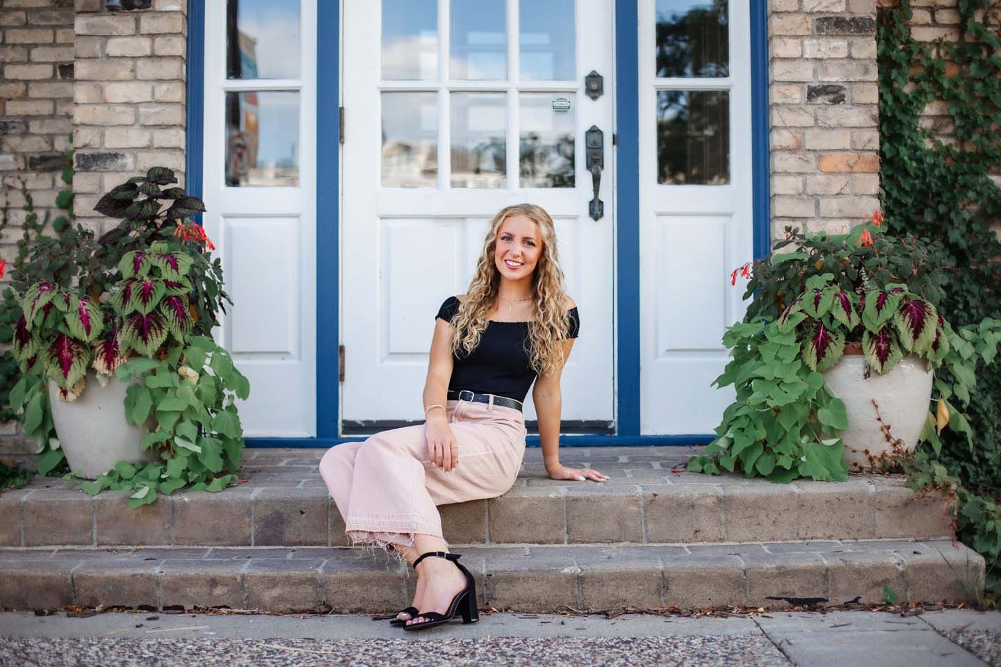 Fashion portrait on brick steps with blue door and greenery featuring pink pants and black top.