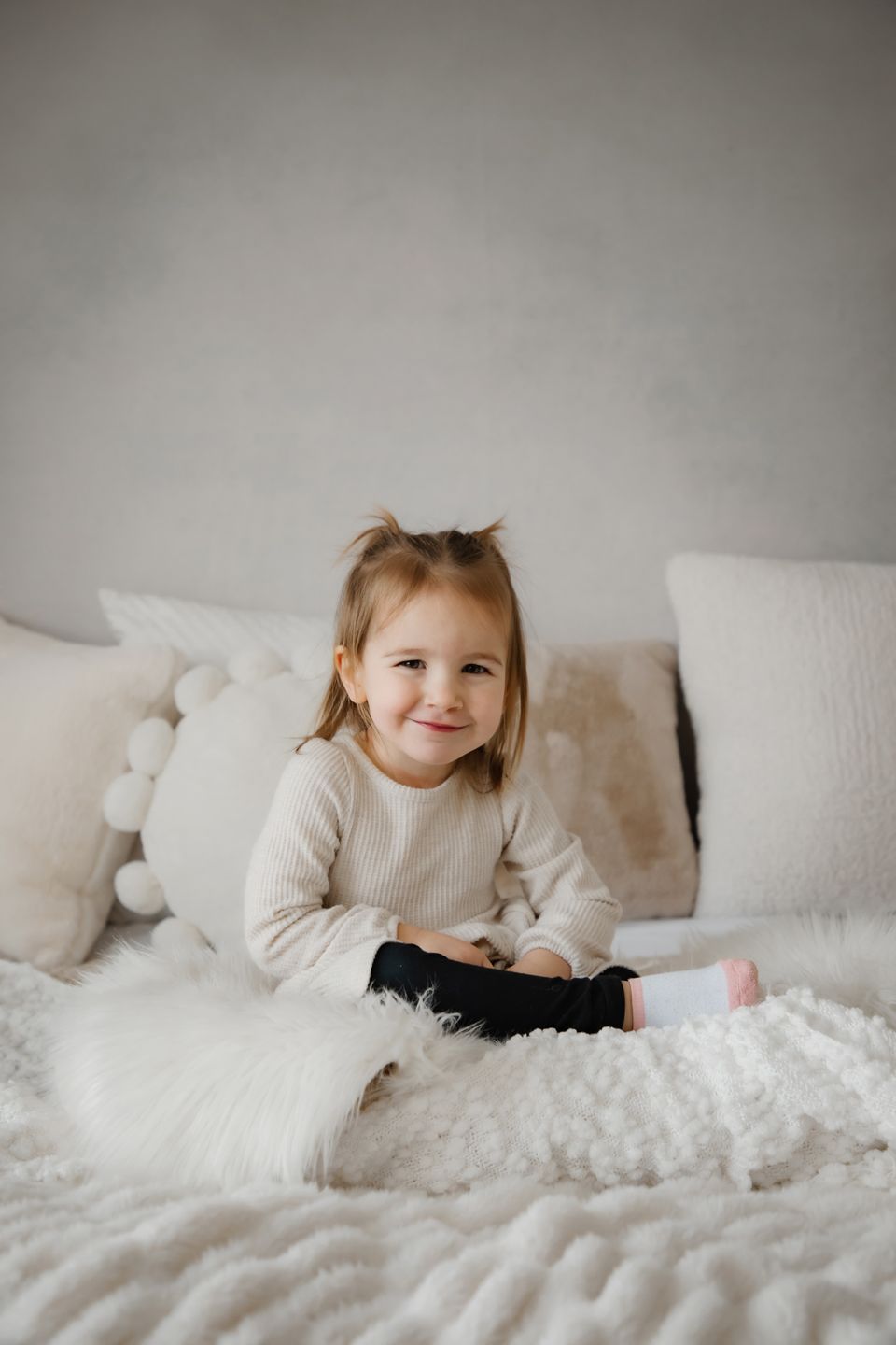 A small child sits smiling on a cozy white bed with plush pillows and blankets.