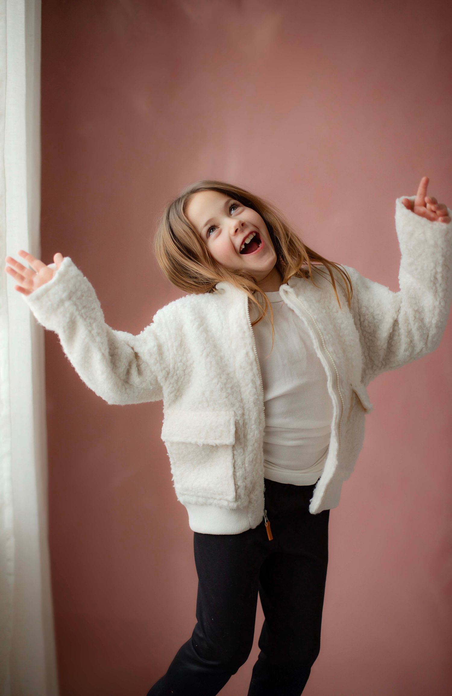 A joyful child wearing a white fuzzy jacket jumps with arms outstretched against a pink studio backdrop.