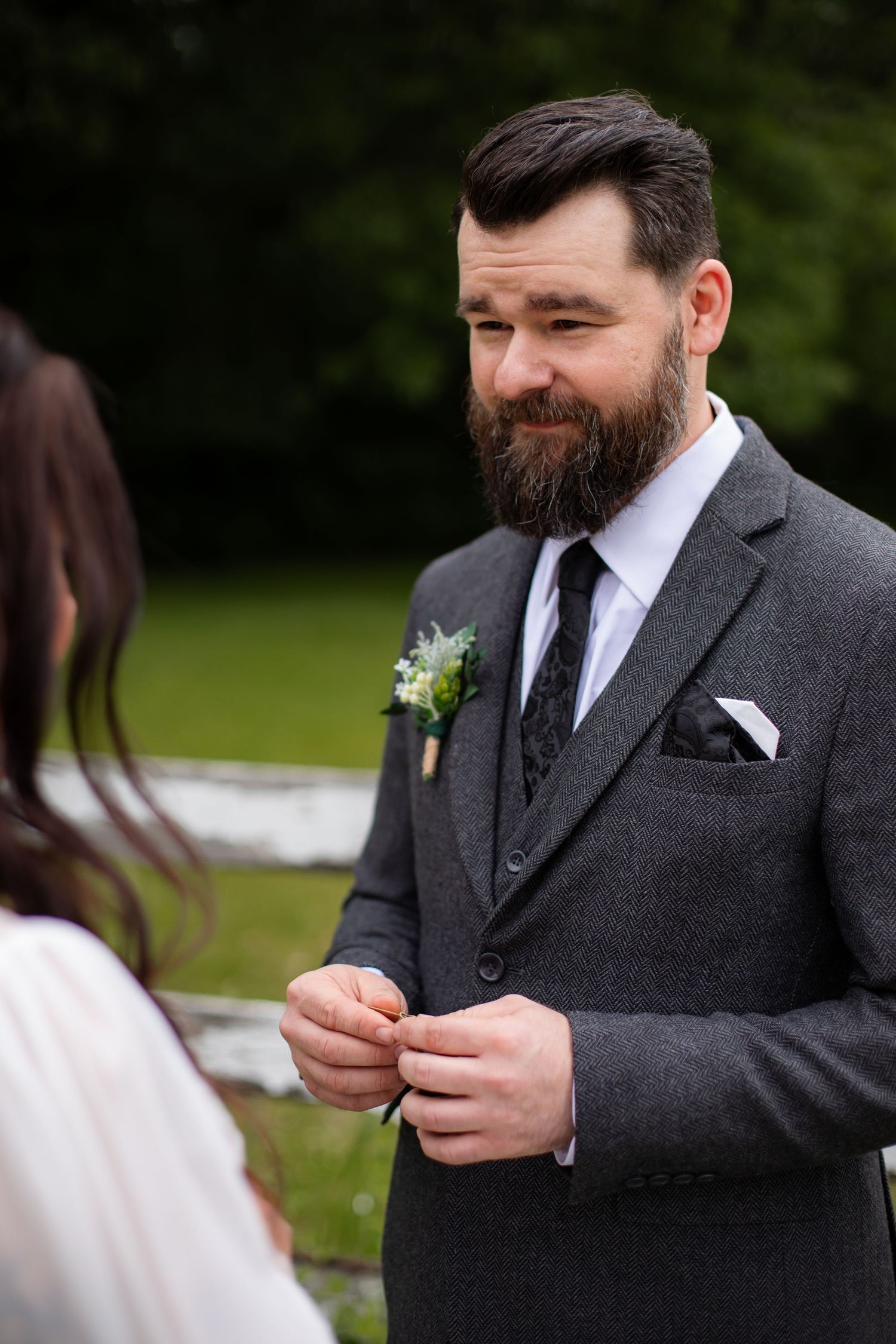 A groom in a dark suit adjusts his boutonniere while standing outdoors in a garden setting.