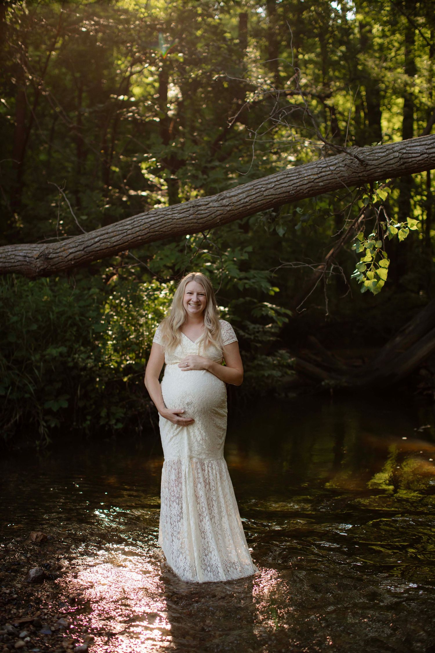 Pregnant woman in white lace gown standing in a sunlit forest creek, cradling her baby bump.