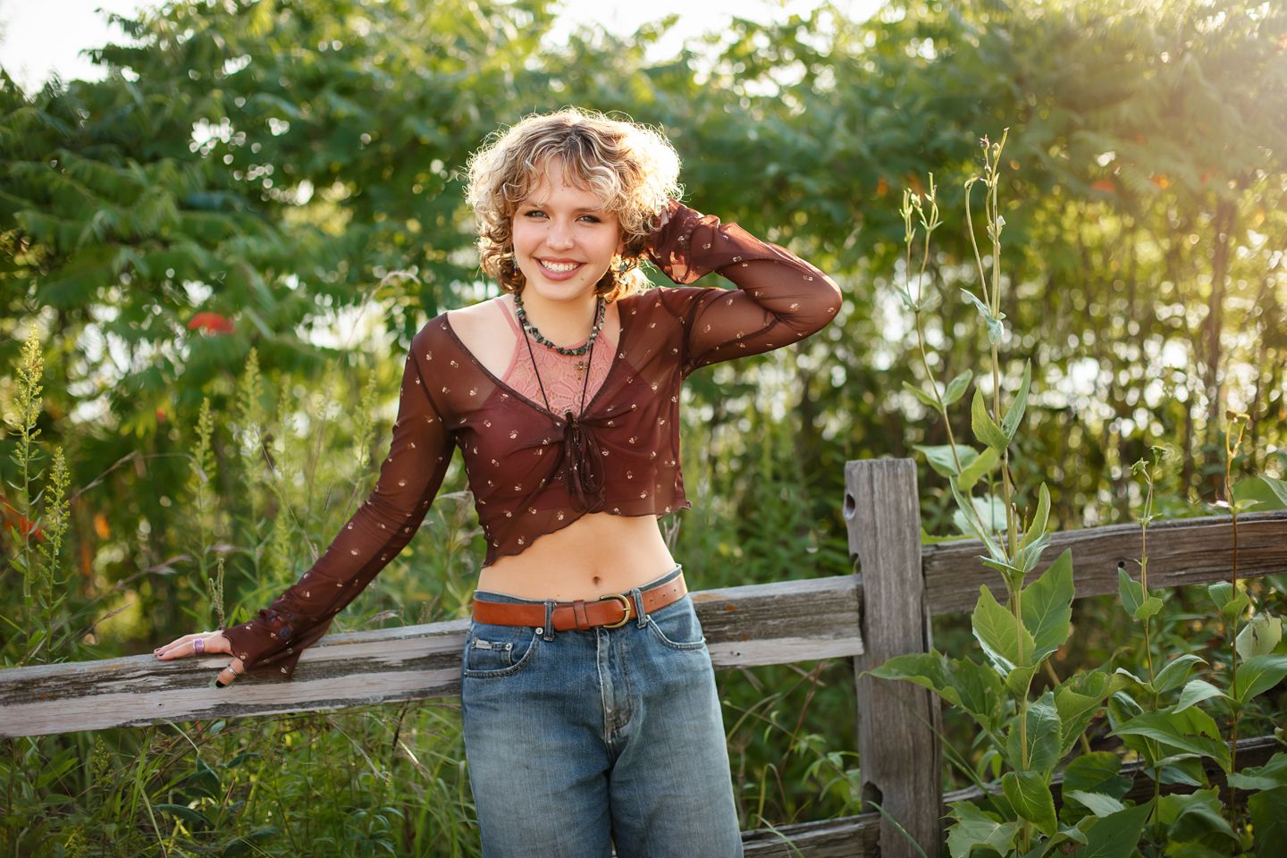 Someone in a burgundy cropped top and jeans leans against a rustic wooden fence at sunset in a garden setting.