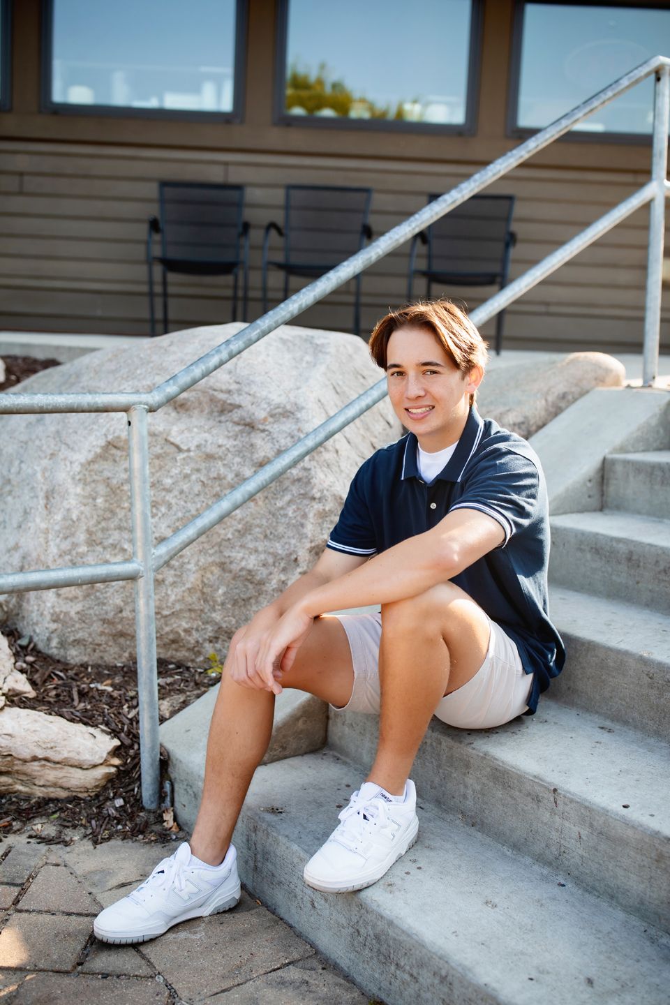 A person in a navy polo shirt and khaki shorts sits casually on concrete steps on a sunny day.