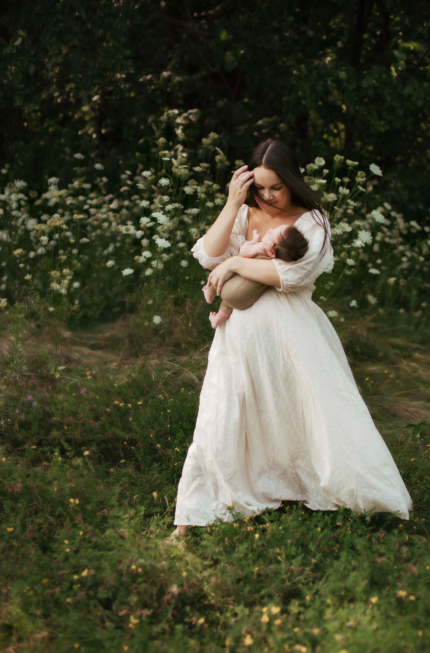 A woman in a white gown holds a newborn baby in a lush green meadow with wildflowers blooming behind her.