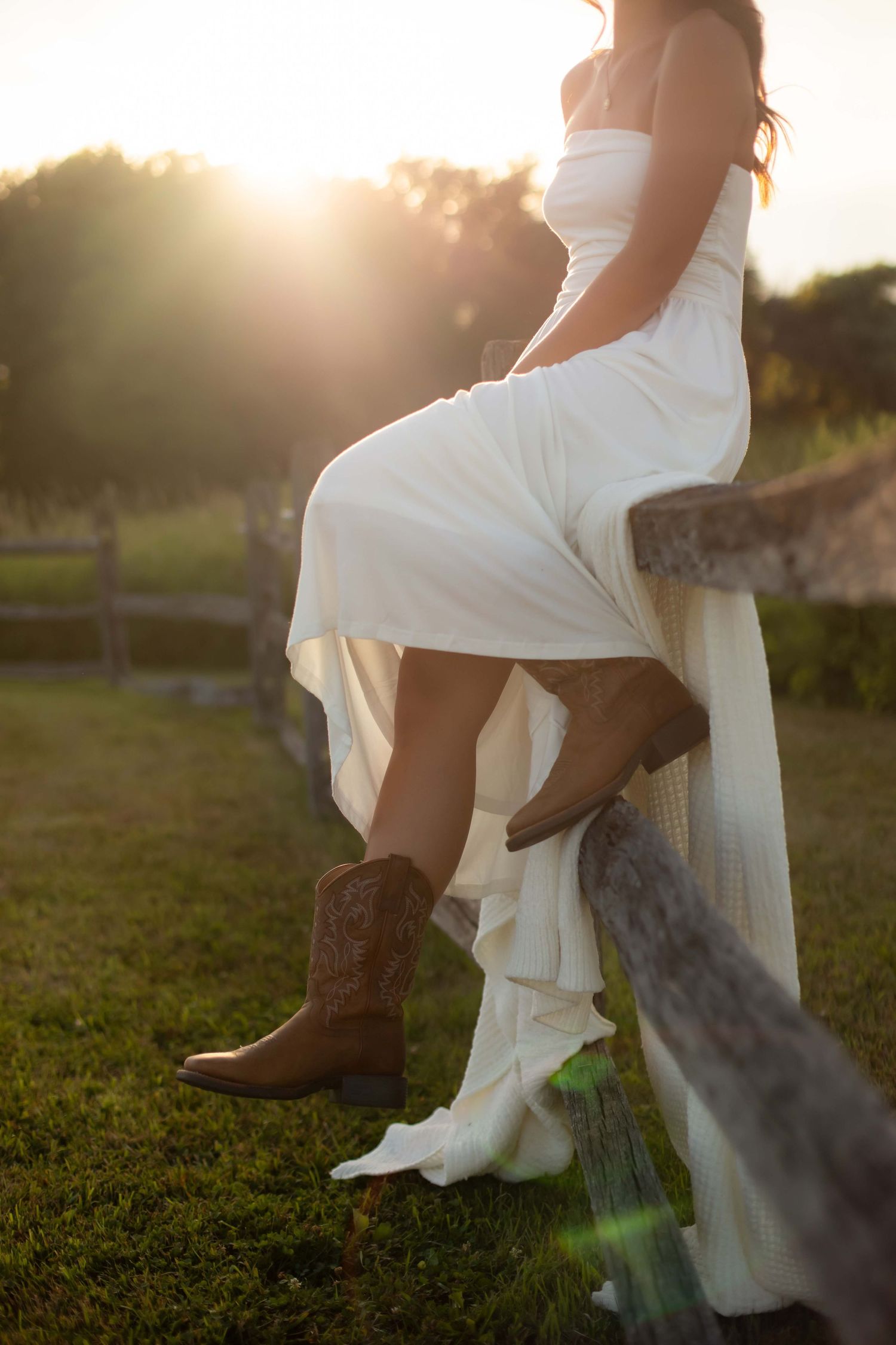 Close-up of woman in white dress wearing brown cowboy boots perched on wooden fence at sunset.