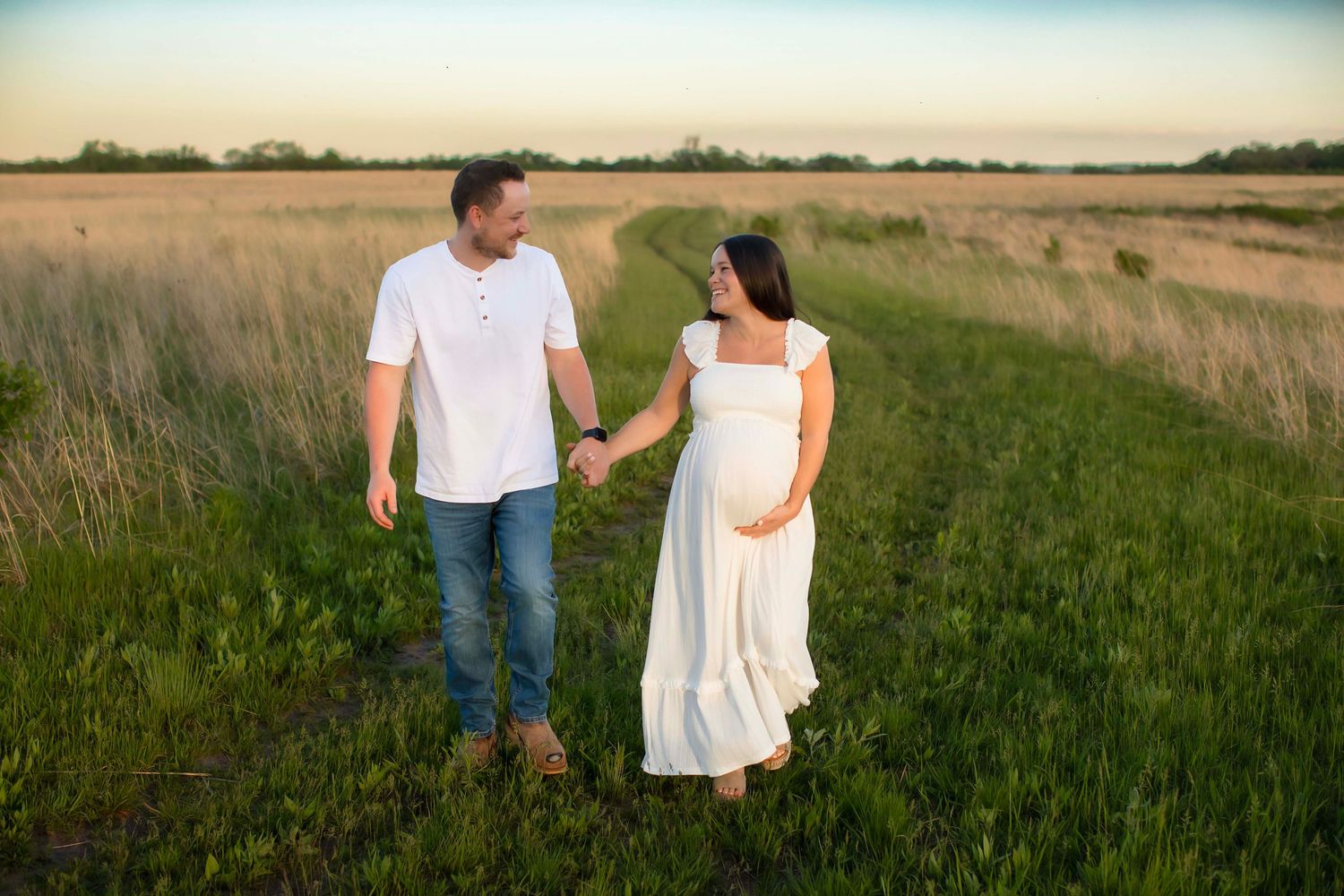Pregnant couple holding hands walking through a green field at sunset, woman in white dress, man in white shirt and jeans.