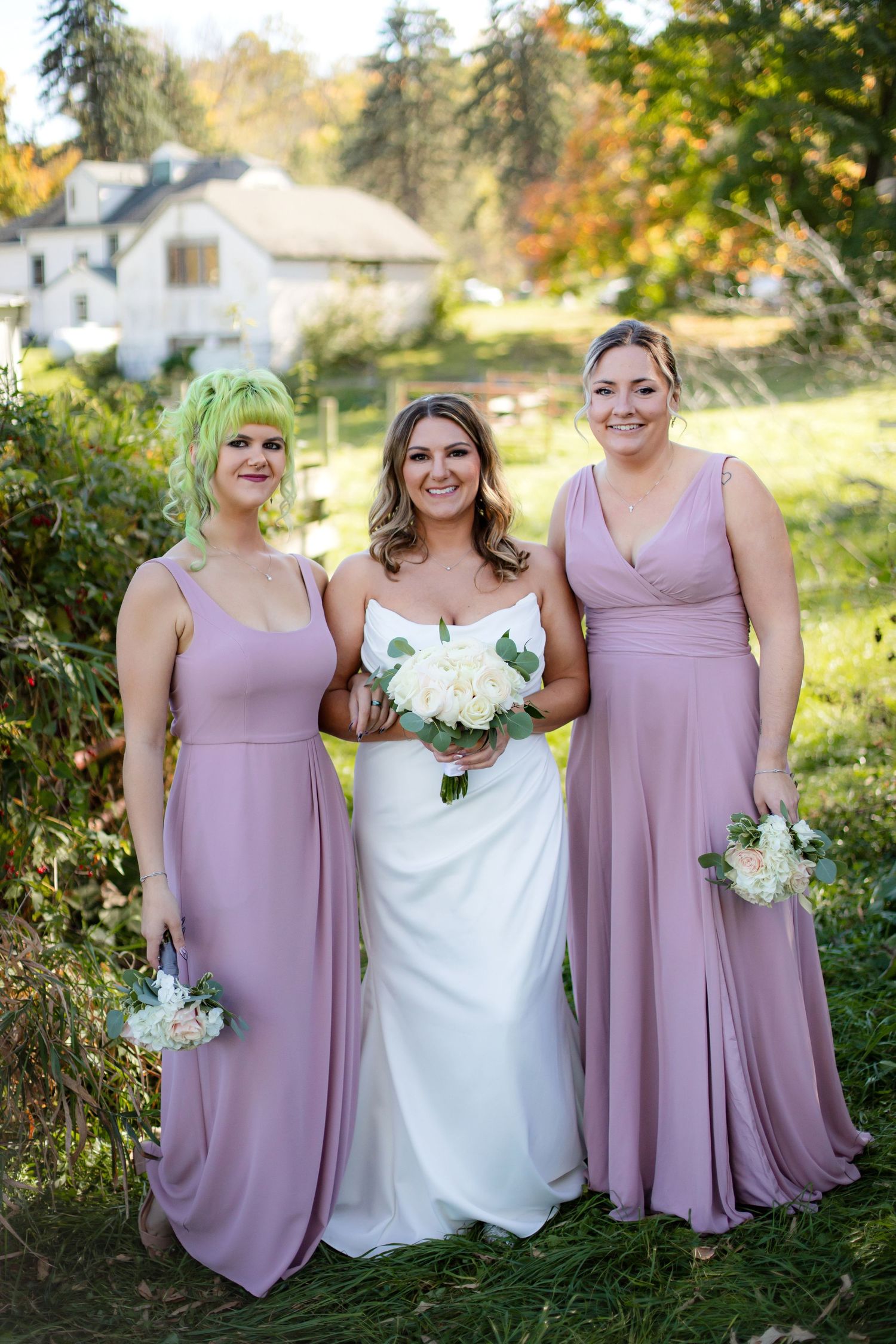 Three women in coordinating mauve dresses pose for a formal wedding photo in a garden setting.