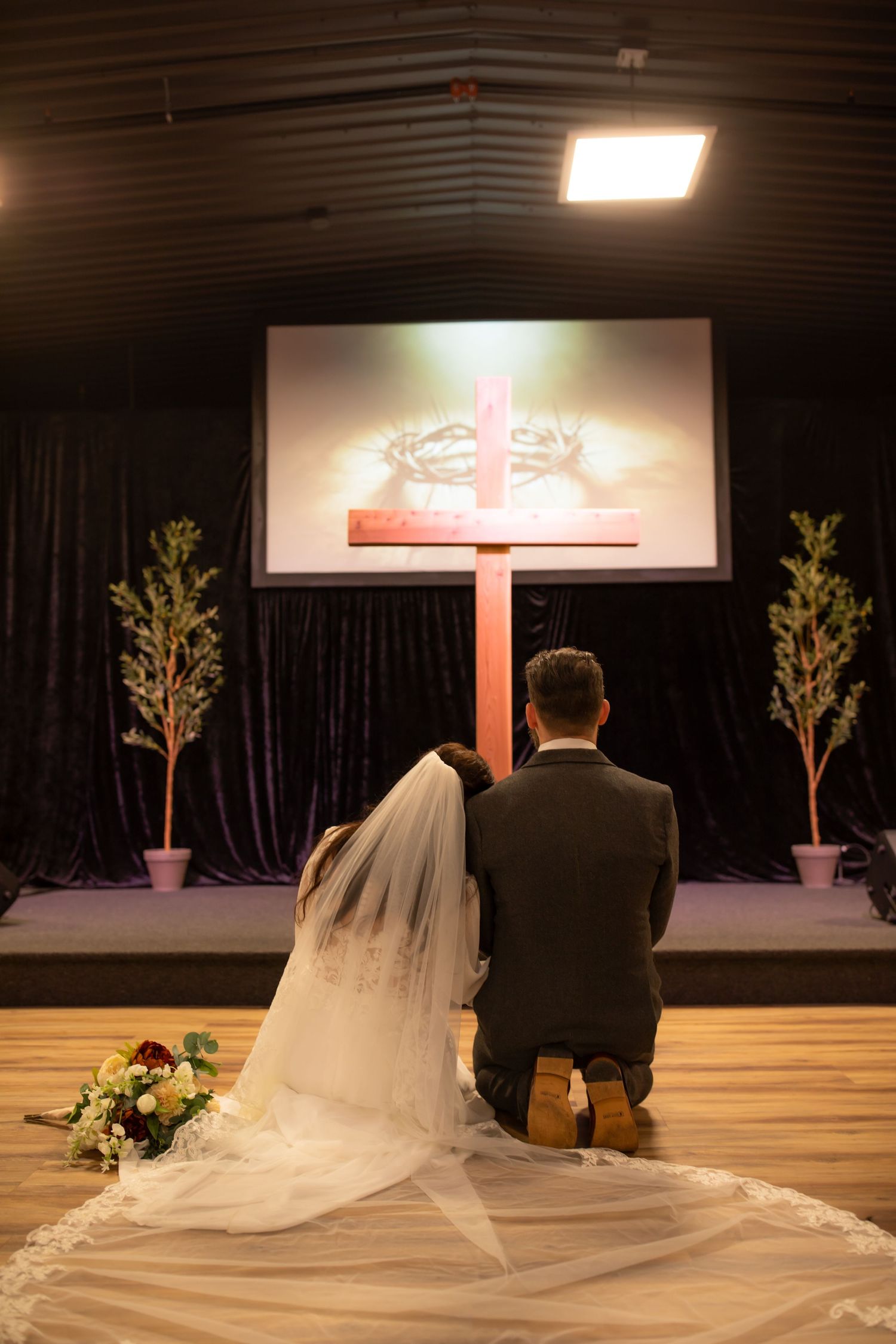 A long flowing white wedding veil trails behind a couple kneeling at a church altar during a ceremony.