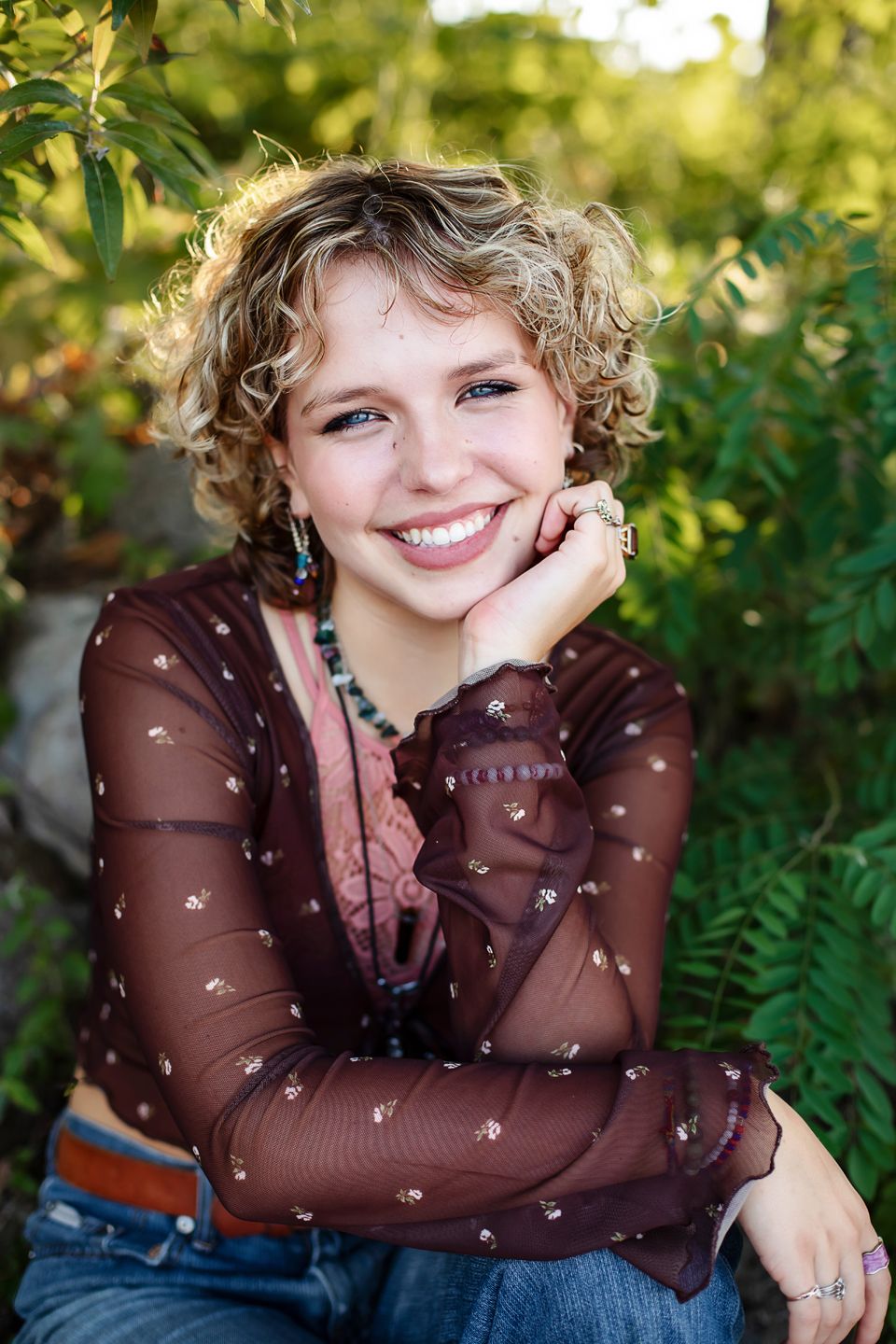 A natural outdoor portrait with curly blonde hair wearing a brown sheer top with small stars in a garden setting.
