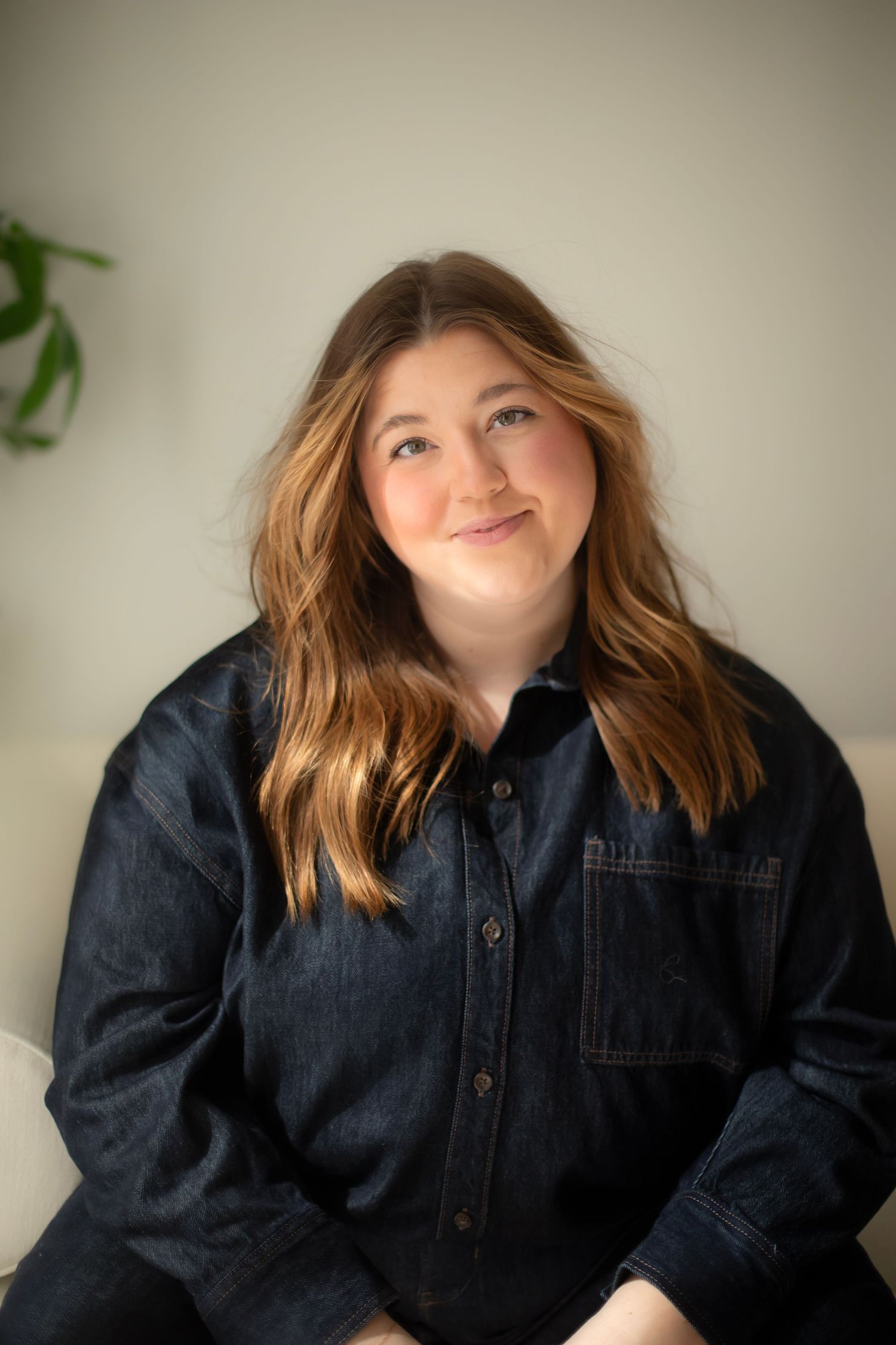 Young woman with auburn hair smiling, wearing a dark denim jacket, seated against a light background.