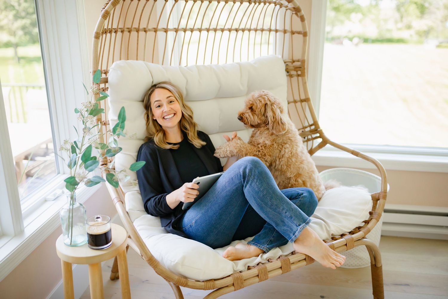 Cozy scene with goldendoodle dog and person relaxing in rattan chair with plush cushions by sunny window.