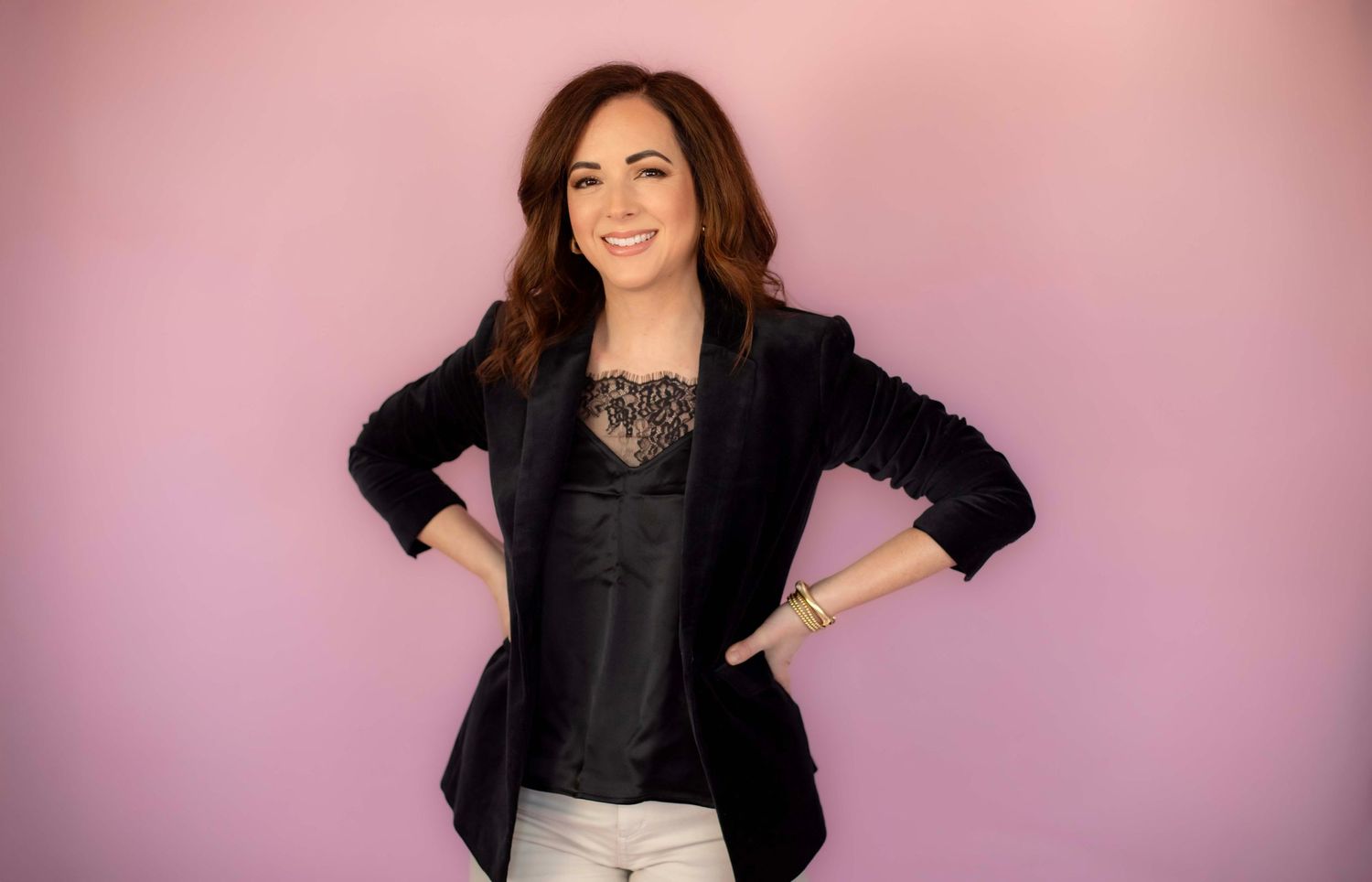 Standing portrait in black velvet blazer with lace top against gradient pink backdrop.