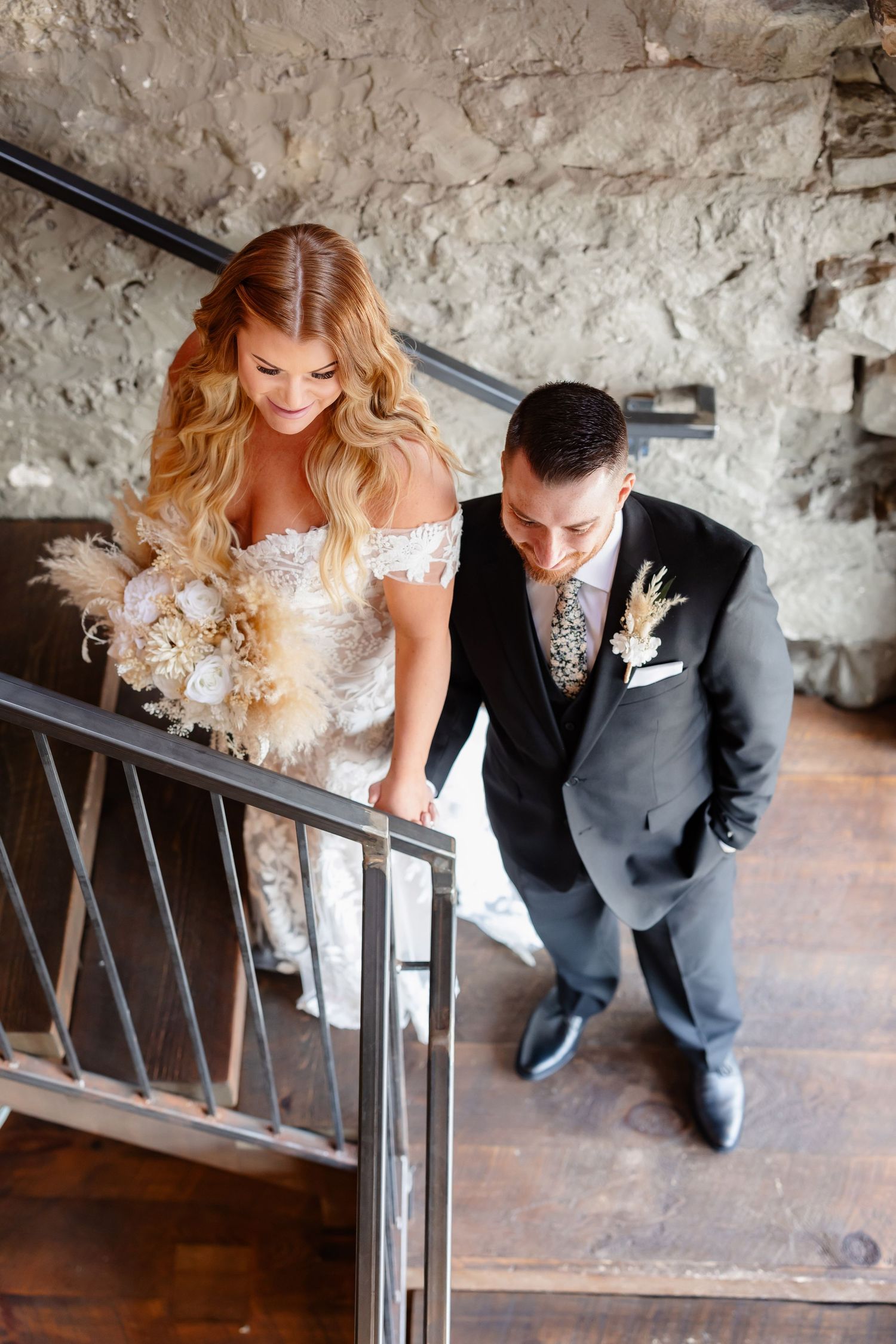 A couple on a rustic staircase with stone walls, the bride wearing an off-shoulder lace gown.