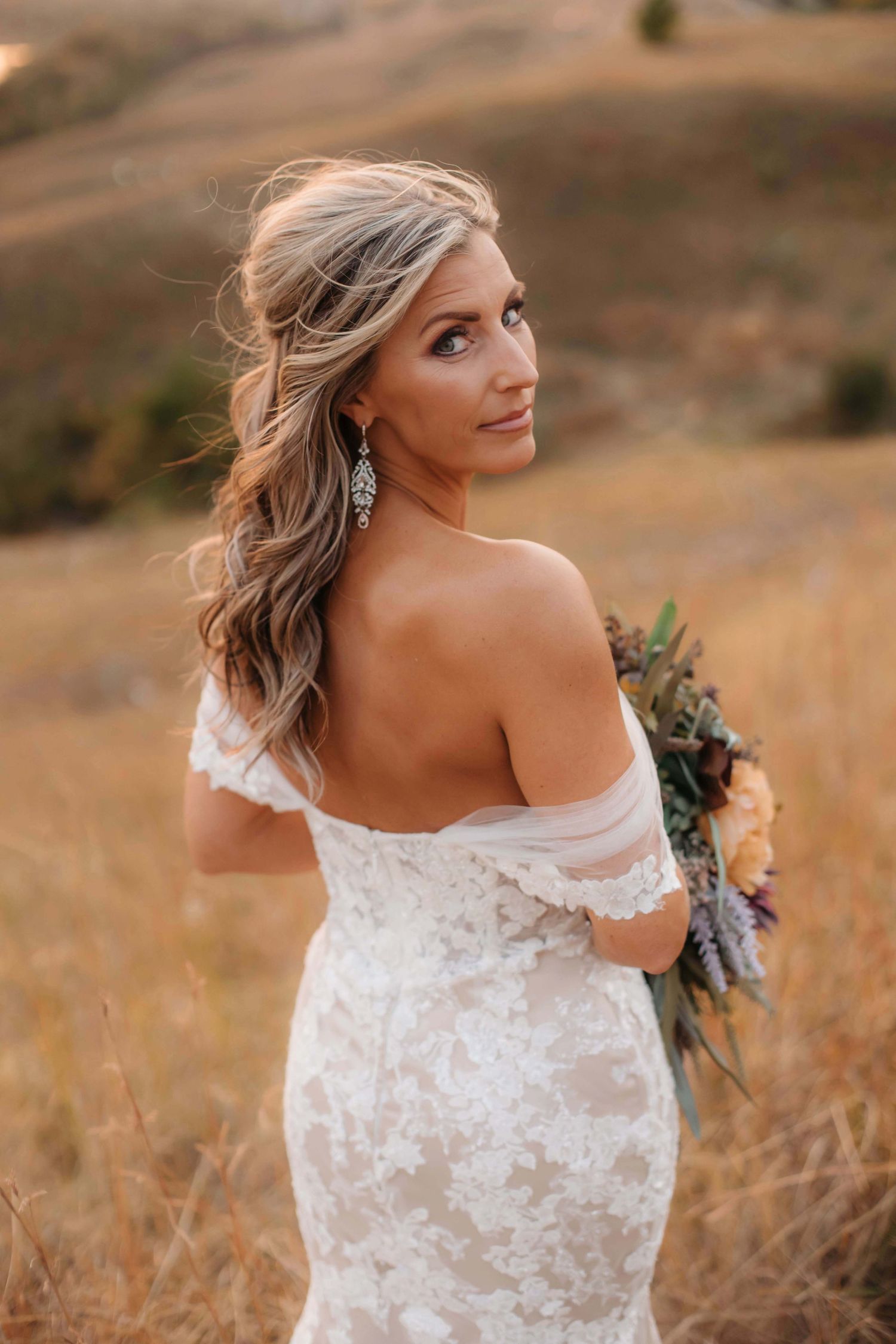 A bride looks over her shoulder while holding a vibrant bouquet against a sunset backdrop.