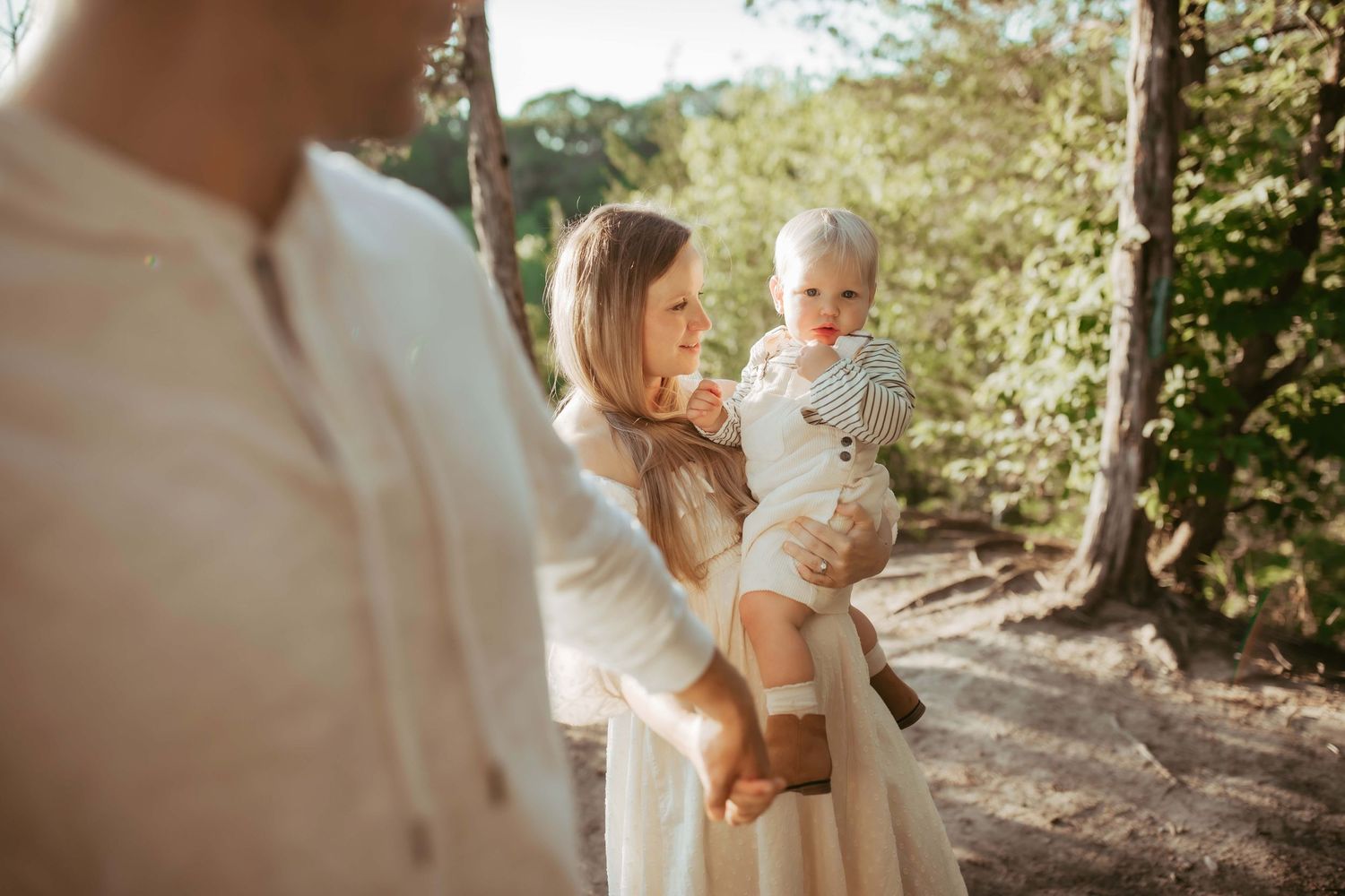 A sunlit outdoor family portrait with a toddler wearing stripes being held in a natural setting.