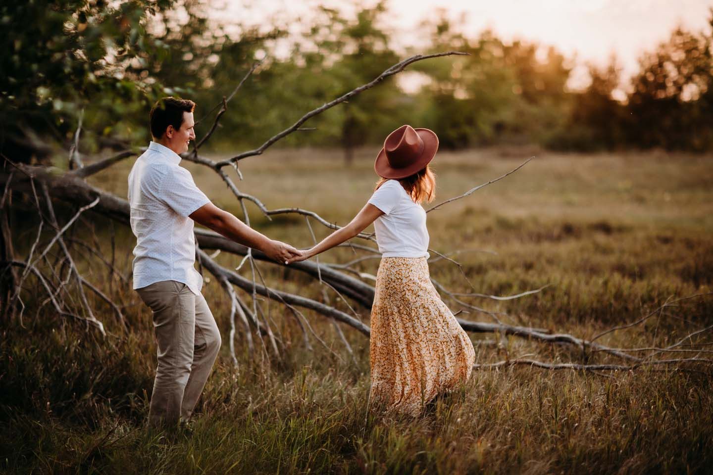 Couple holding hands and dancing in a golden field at sunset with fallen trees and tall grass in the background.