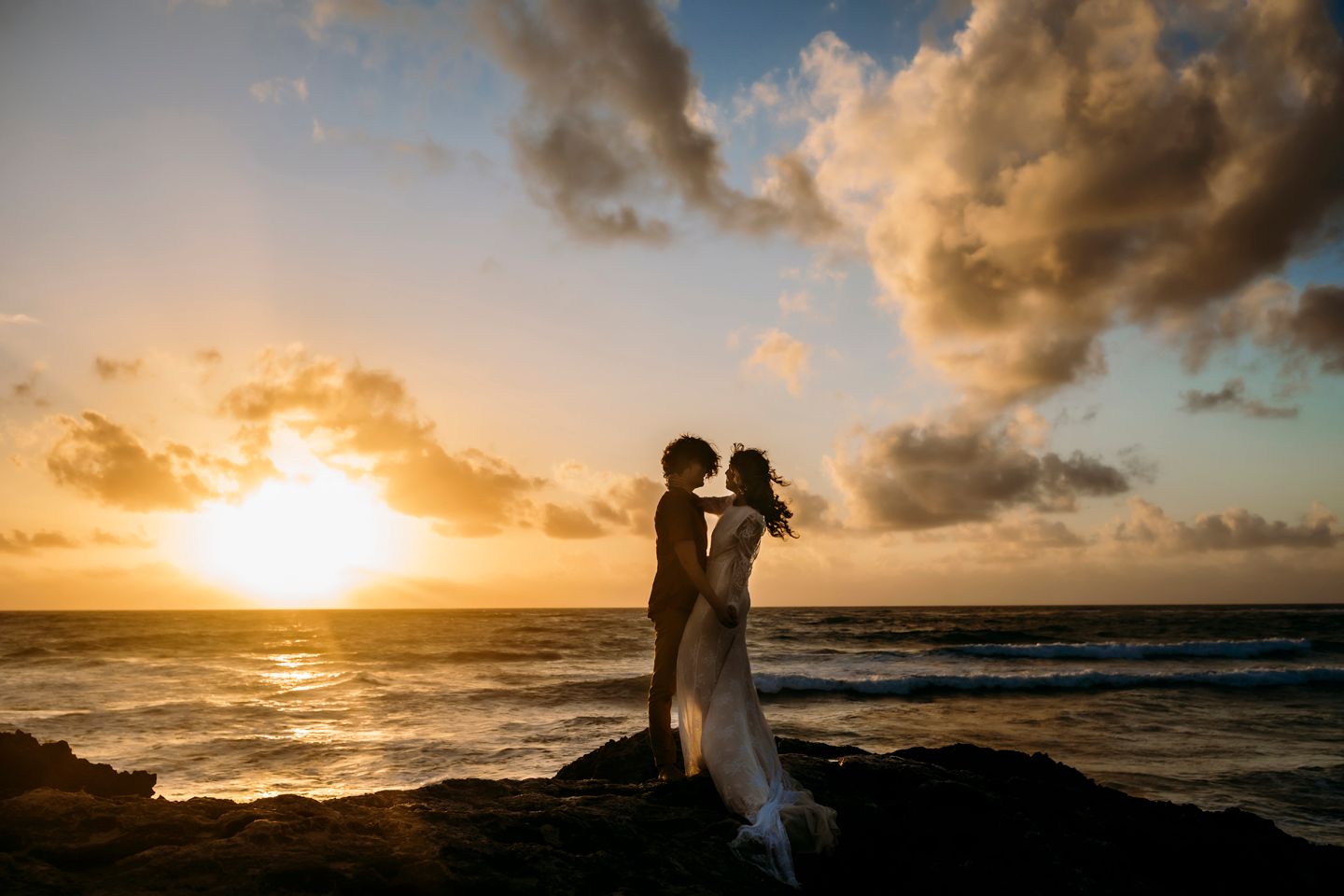 Dramatic silhouette against golden sunset sky with crashing waves on rocky shore.
